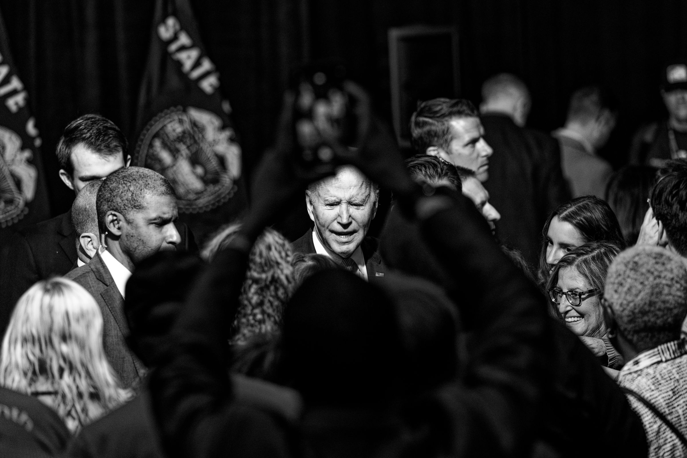 A high-contrast black and white photograph capturing a crowded political event. President Joe Biden's face is framed by the raised hands of a supporter taking a cell phone picture, surrounded by a group of supporters and Secret Service.