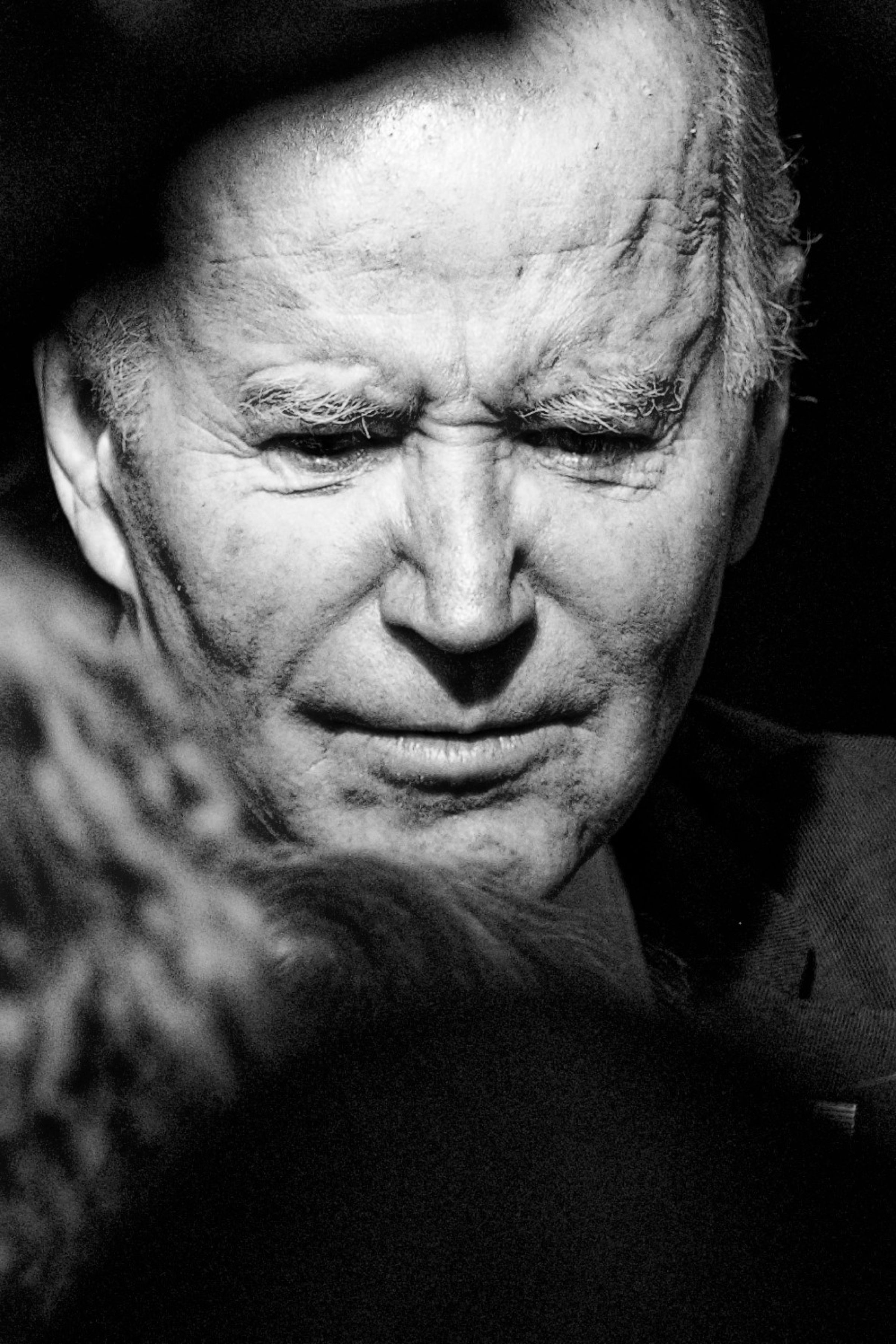 A stark black and white close-up portrait of President Joe Biden with a deeply furrowed brow and intense expression. The high-contrast lighting emphasizes the President Biden's facial features and wrinkles, creating a powerful and emotive image.