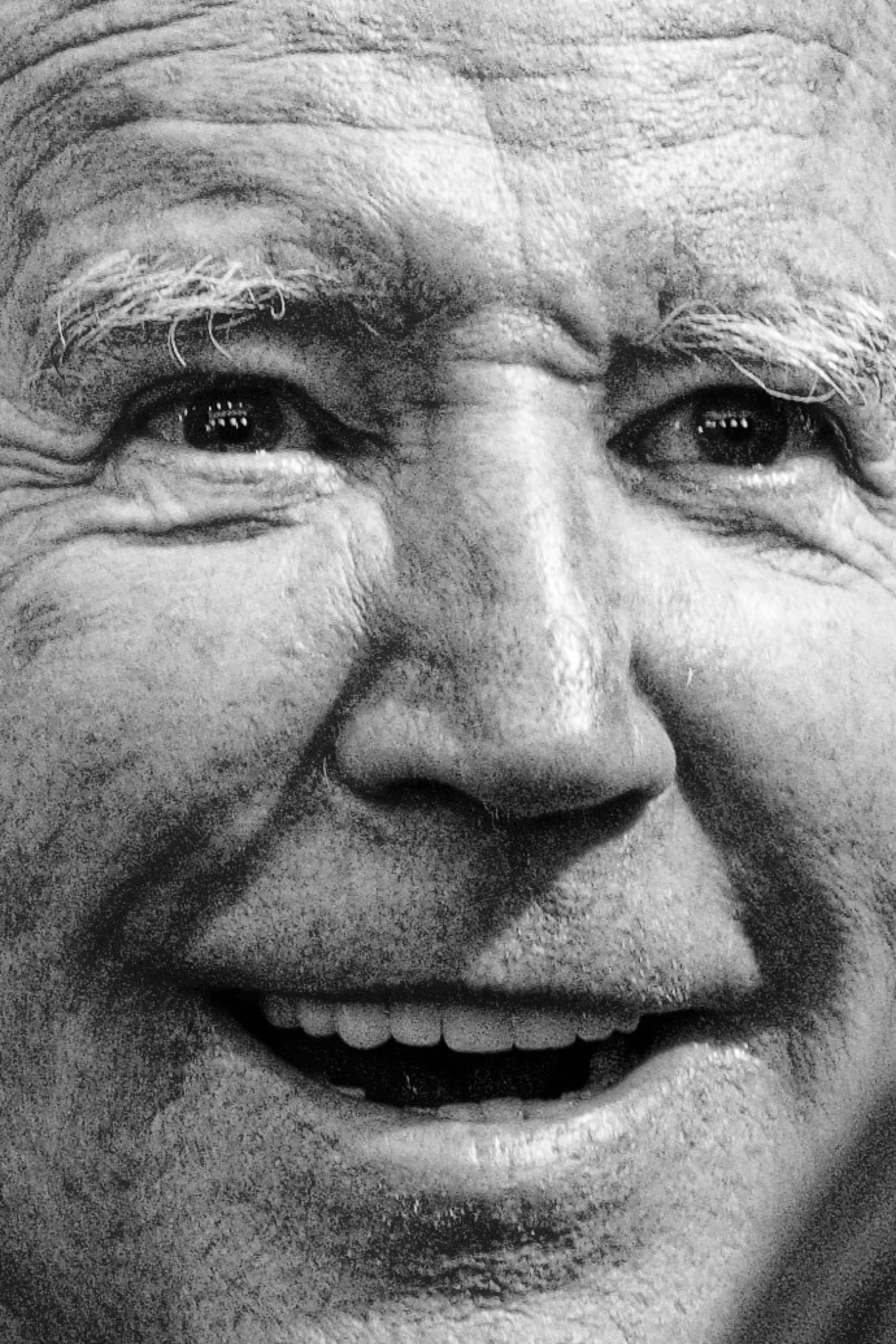 A close-up black and white photograph of President Joe Biden's smiling eyes and face. His mouth is partially open, and the light is reflecting in his eyes.