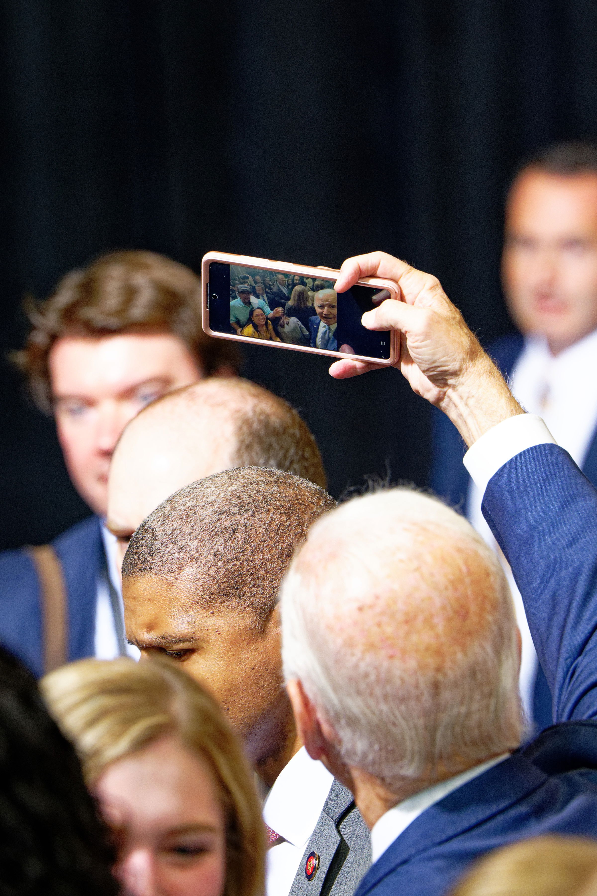 A colorful scene at a political event, with a hand holding up a smartphone to take a photo. The phone's screen reveals President Joe Biden posing with several supporters while a crowded gathering of people in formal attire mingle in the foreground.