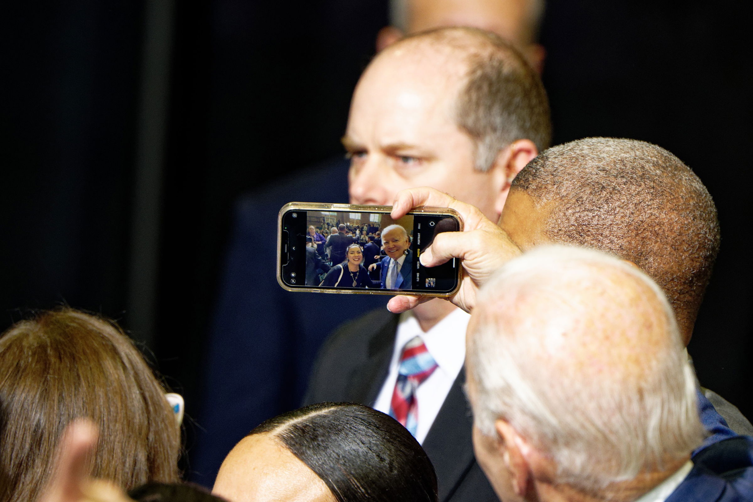 A close-up view of a political event, showing hands holding up a smartphone to capture a photo. The phone's screen reveals President Joe Biden and a supporter smiling for the camera with a crowded room of attendees behind them.