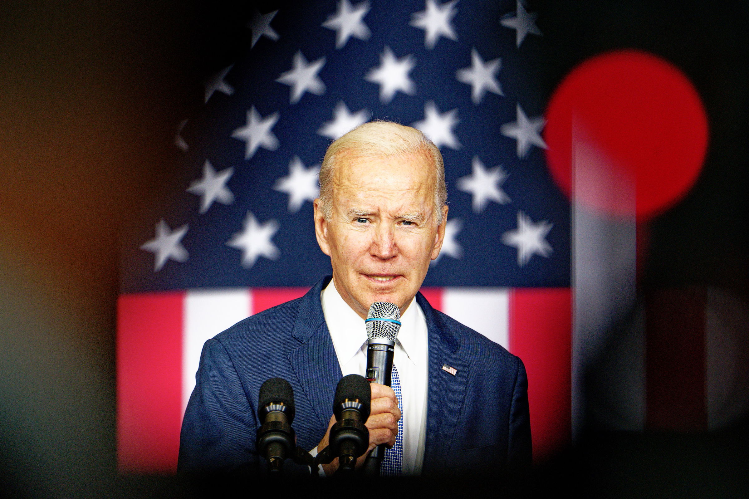 A vibrant color photo of President Joe Biden speaking at a podium with an American flag backdrop. He wears a navy suit and holds a microphone, addressing an audience with a serious expression while looking directly at the camera.