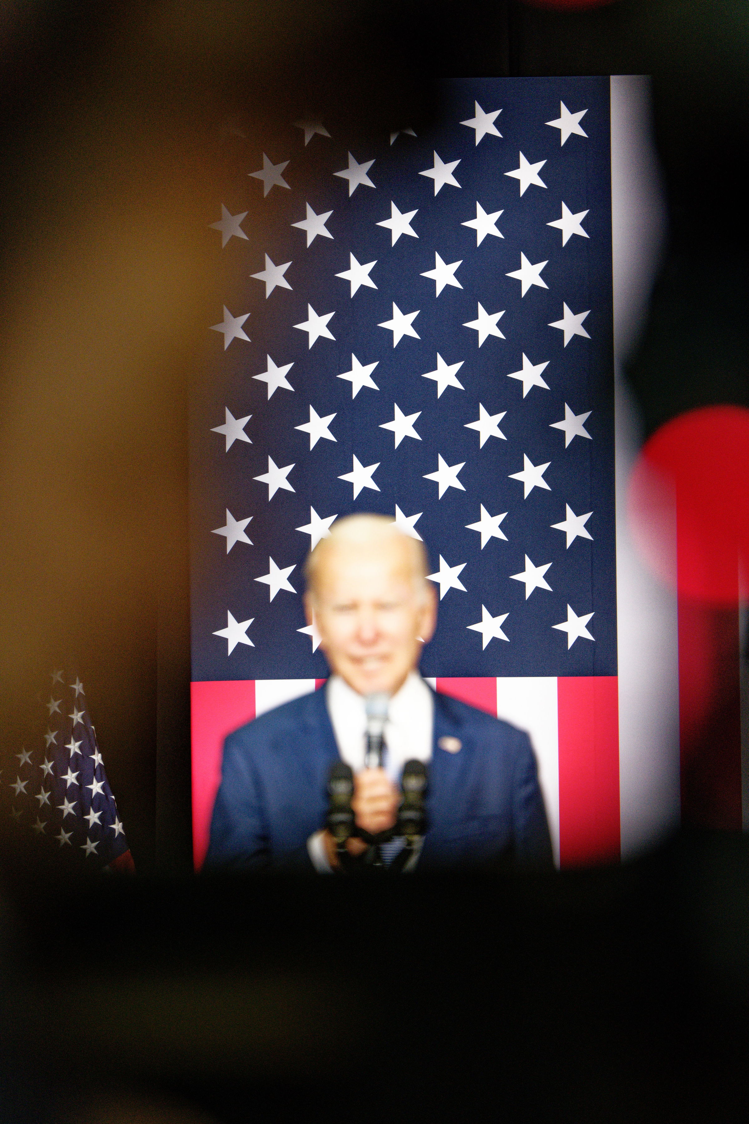 A blured image of President Joe Biden speaking at a podium in the foreground with a sharp American flag in-focus in the background behind him.