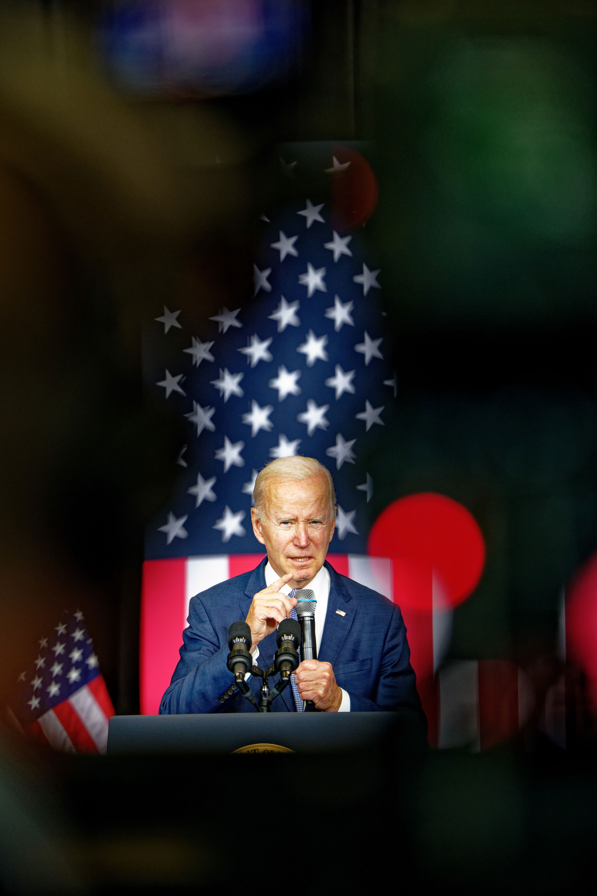 President Joe Biden gesturing while speaking at a podium, framed by the American flag and colorful lens flares. The image captures him mid-speech with an emphatic expression, his finger raised while looking directly into the camera.