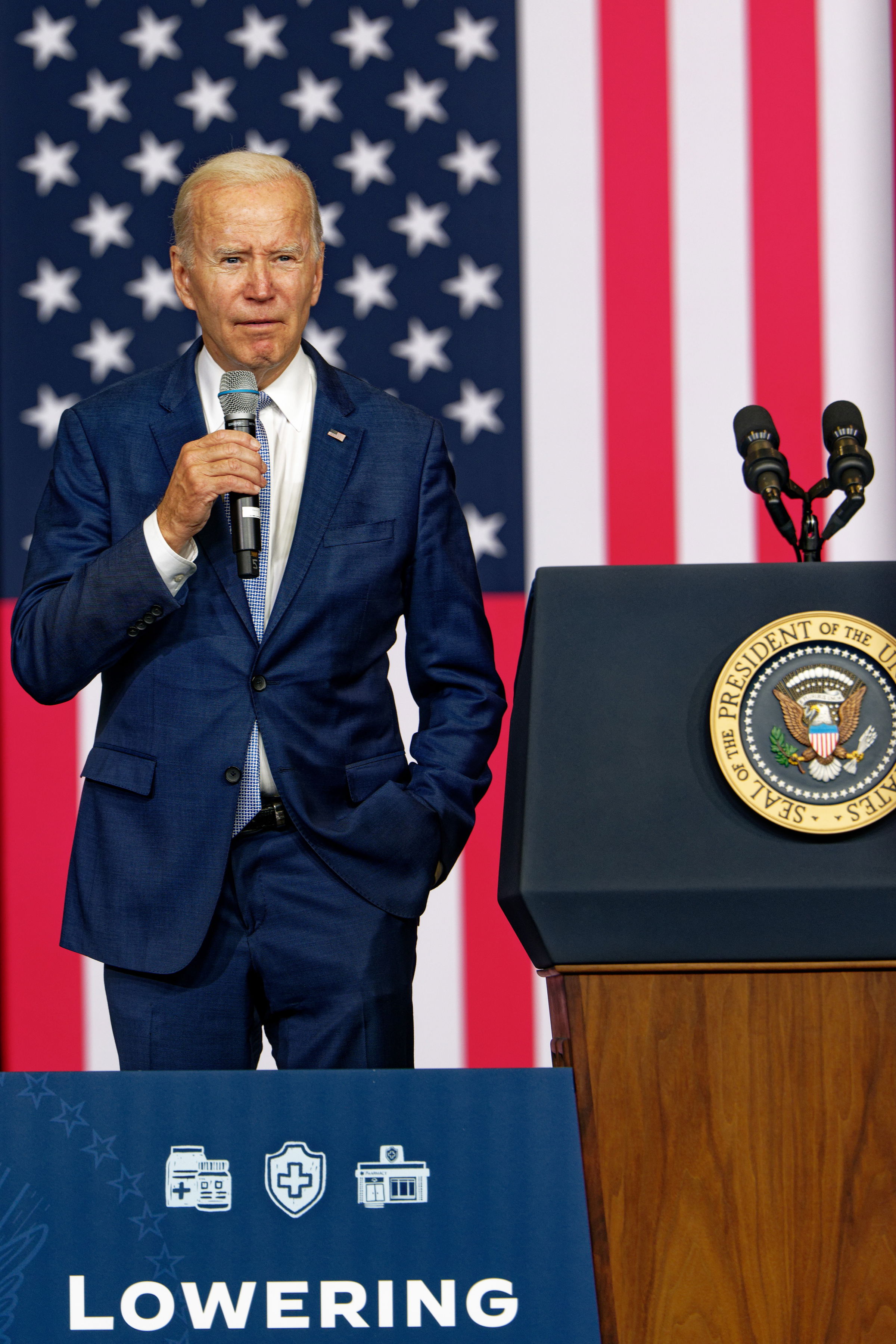 President Joe Biden speaking at a podium with the presidential seal. He wears a dark blue suit and gestures with one hand. The backdrop features the American flag, and a sign that reads "LOWERING" is visible in the foreground.