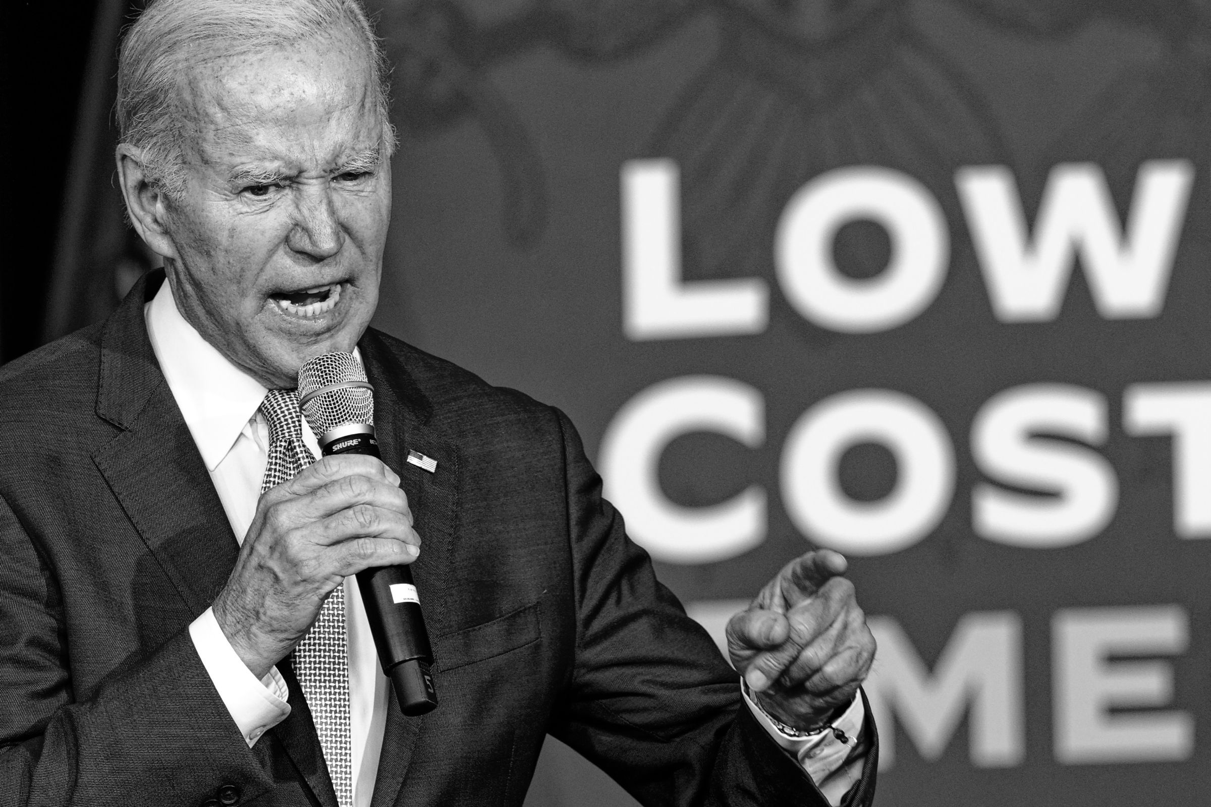 President Joe Biden speaking passionately at a podium, gesturing with one hand while holding a microphone. He wears a dark suit and tie. Behind him, a sign partially visible reads "LOW COST". The image is in black and white, emphasizing Biden's emphatic—angry, even—expression.