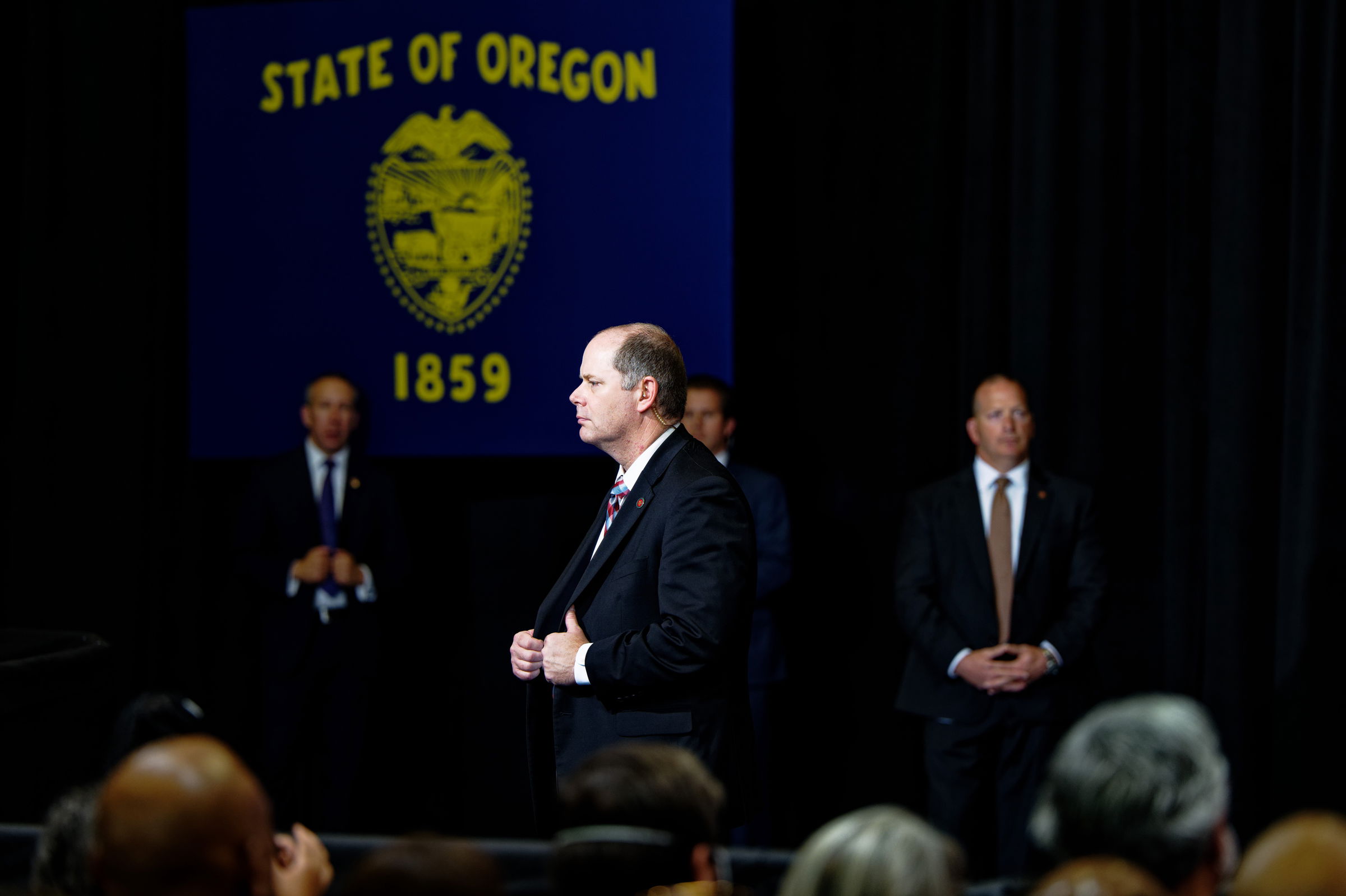 Three Secret Service agents in suits stand on a stage at a political event with the State of Oregon flag prominently displayed behind them. A fourth agent stands at the forefront, his hands gripping his suit jacket, while the others flank him on either side. The audience is visible in the foreground.