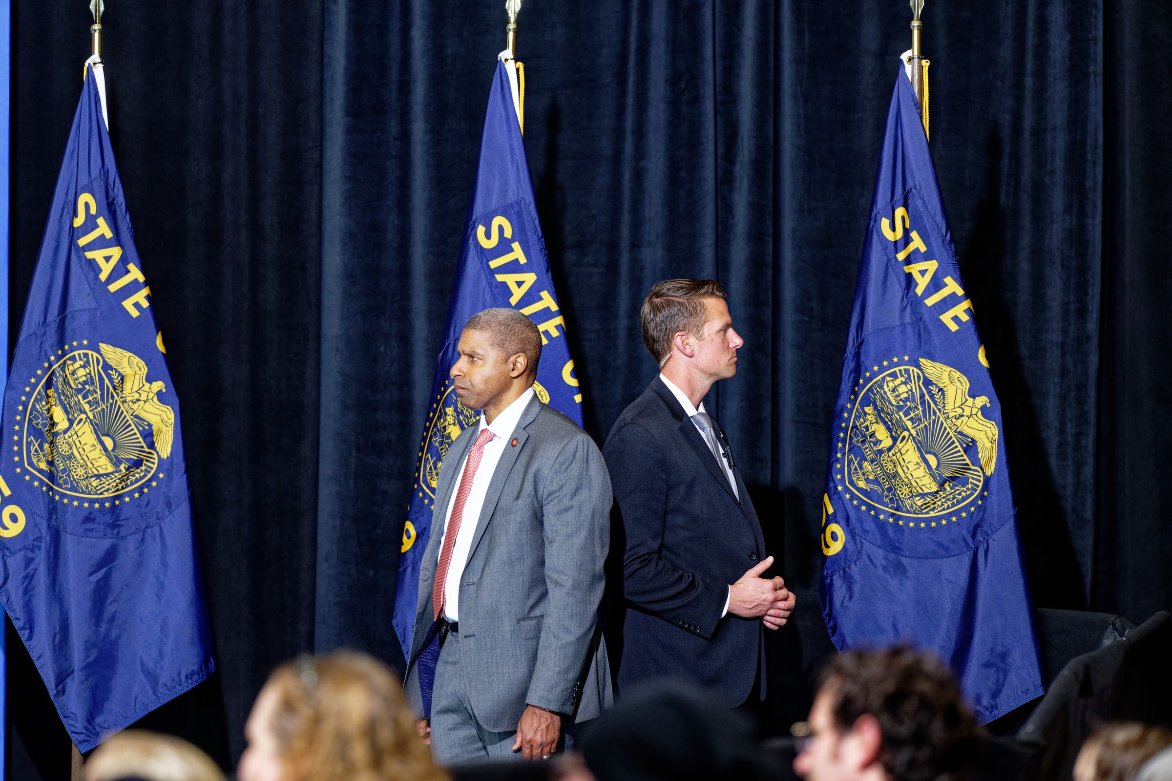 Two Secret Service agents in suits stand in front of three large State of Oregon flags. One man, wearing a gray suit with a red tie, stands slightly in front of the other, who wears a dark suit. The audience is visible in the foreground.