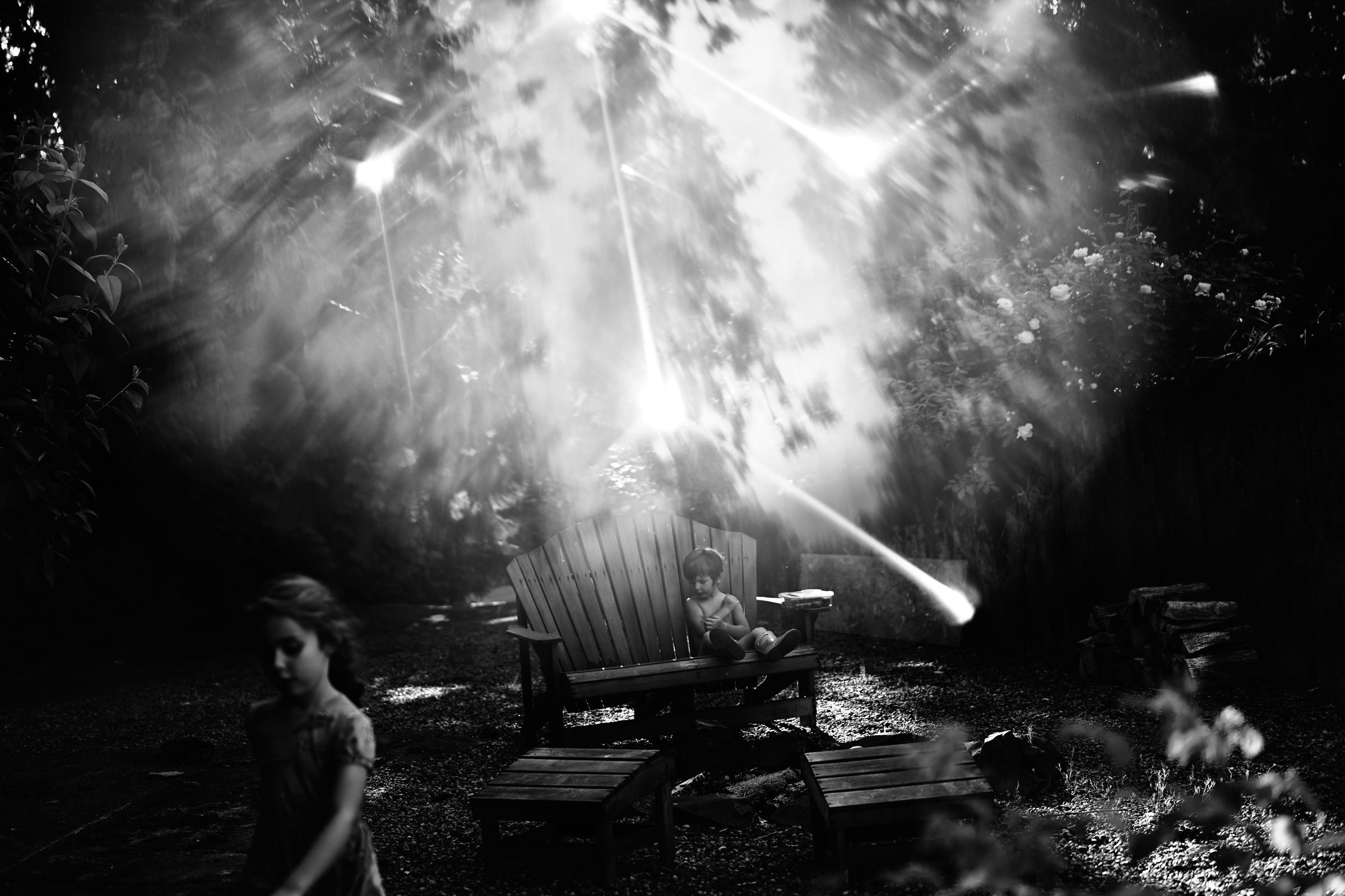 A black and white photo of children in the backyard at dusk. A boy sits on an Adirondack chair and a girl walks nearby. Bright light refracts across the scene in a geometric pattern.