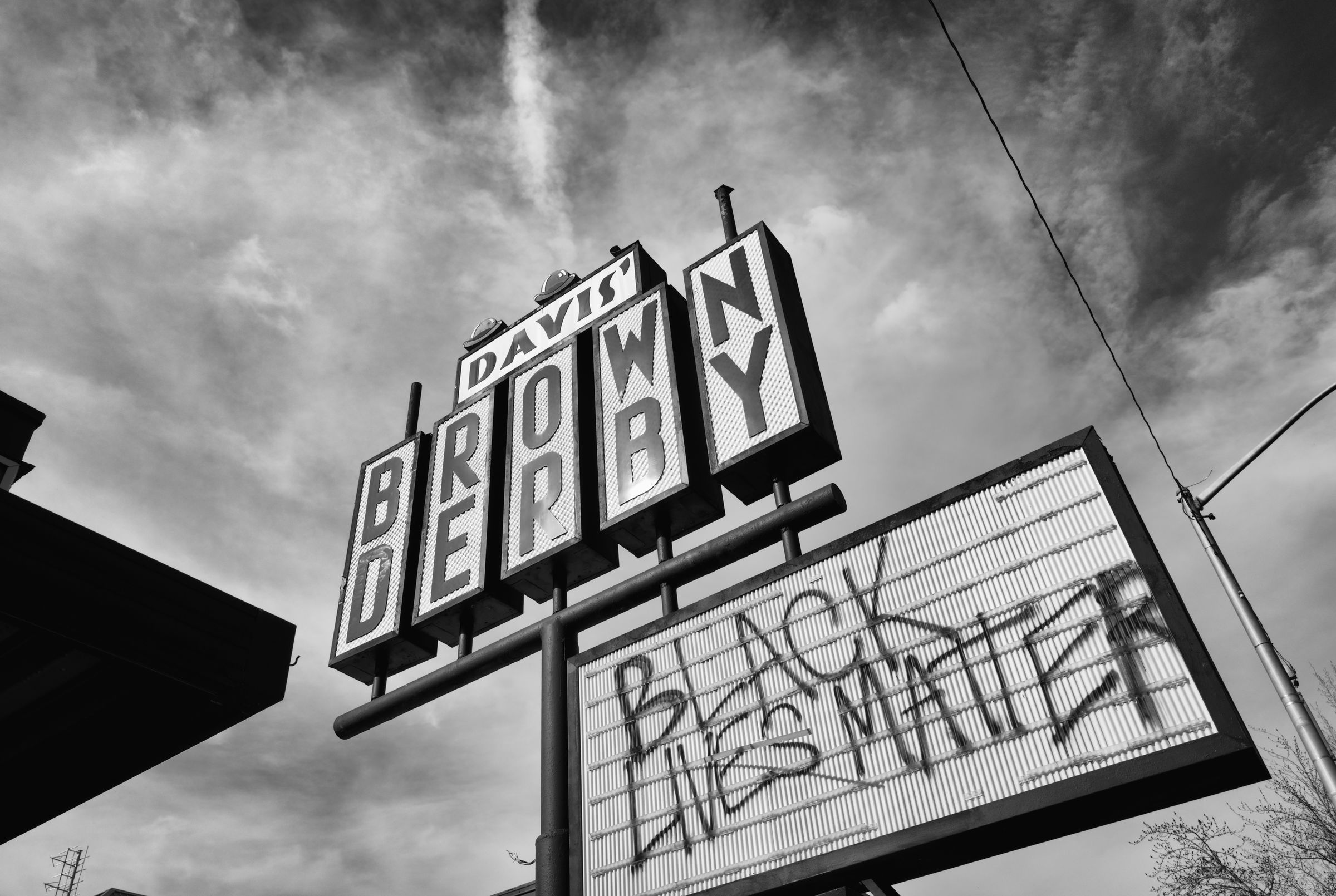 Vintage neon sign for Davis' Brown Derby restaurant against a cloudy sky, with a separate spray painted sign reading "Black Lives Matter".