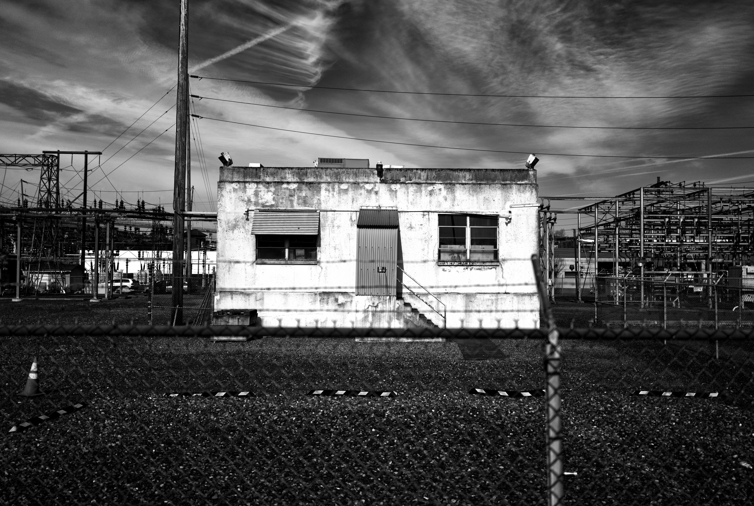 A weathered white building in an industrial area, surrounded by electrical equipment and a chain-link fence, under dramatic cloudy sky.