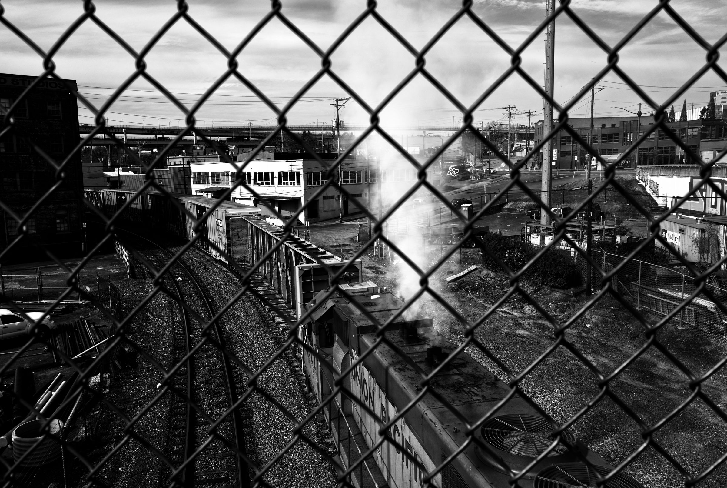 Urban railway yard viewed through chain-link fence, with tracks, buildings, and steam rising in a black and white industrial landscape.