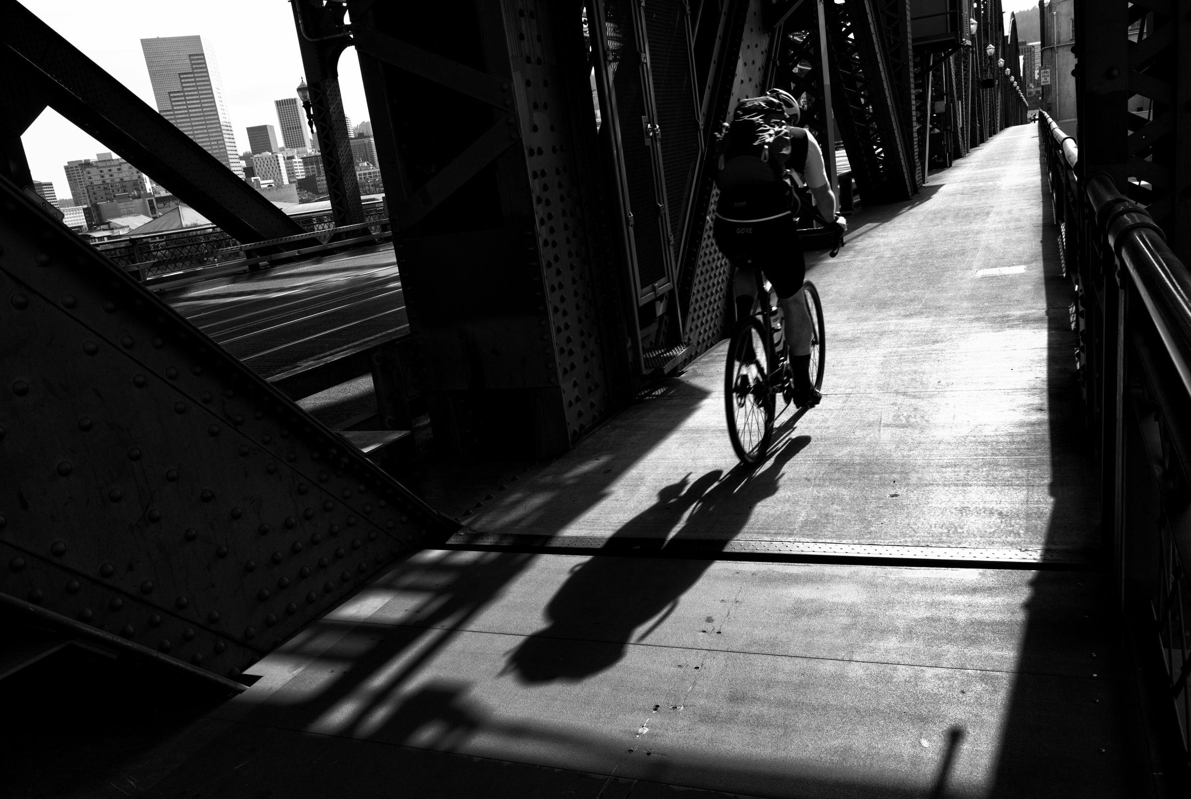 A cyclist crosses an urban bridge, casting long shadow amid steel girders with city skyline visible in background.