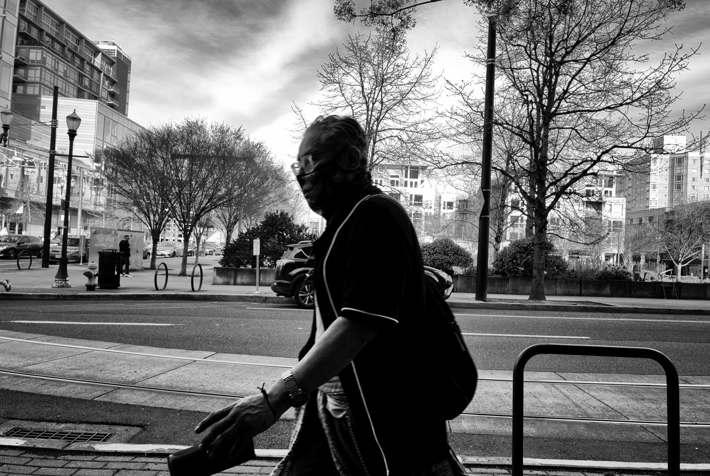 A person in dark clothing passing on a sidewalk, surrounded by trees and modern buildings.