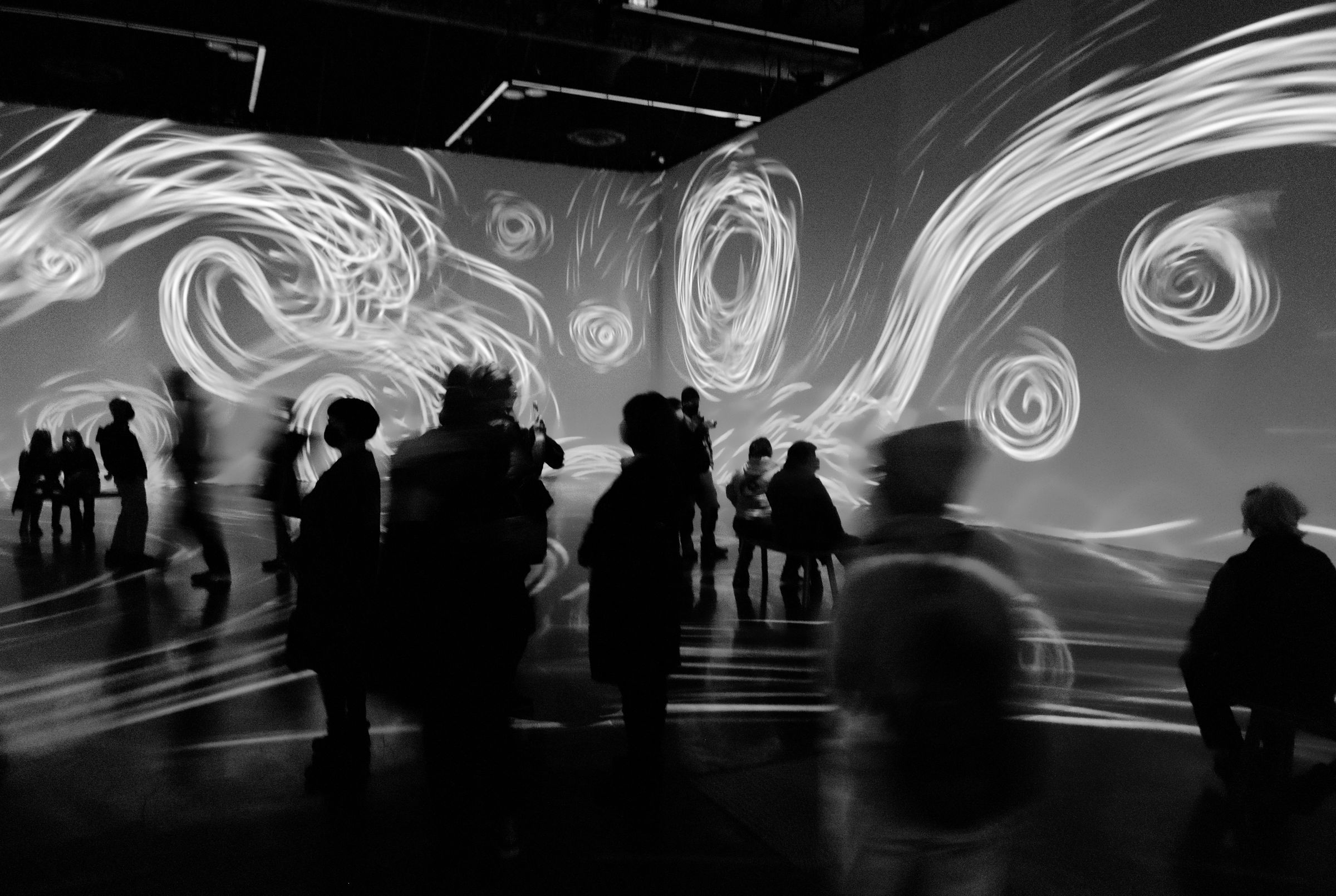 Silhouettes of people viewing large-scale abstract light projection in a dark room, with swirling patterns creating an immersive art experience.