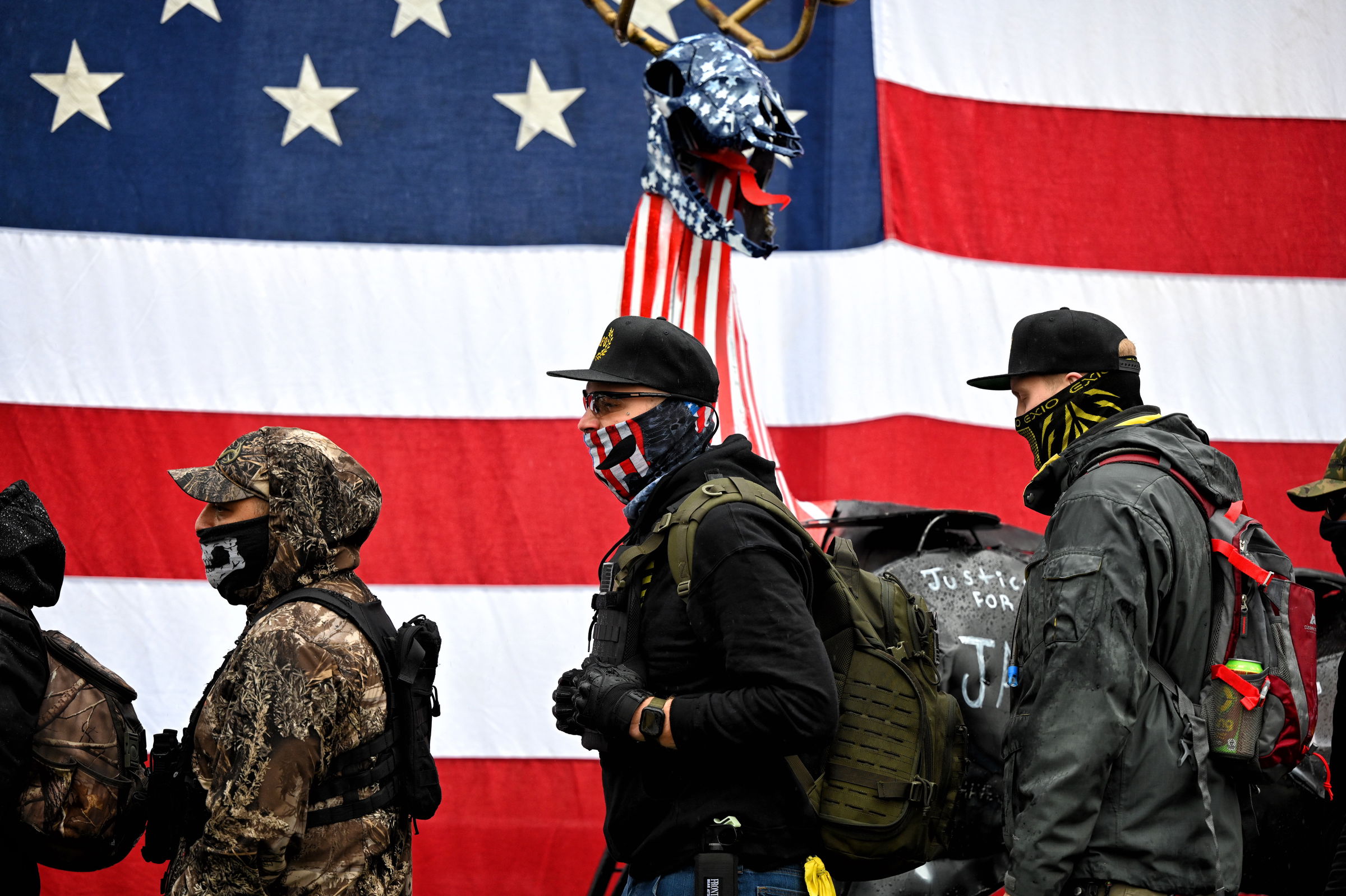 Armed protesters in tactical gear and face masks stand in a line before a large American flag and a deer-like sculpture, with one wearing a camouflage hat and another carrying a backpack with the partially obscured text "Justice for J".