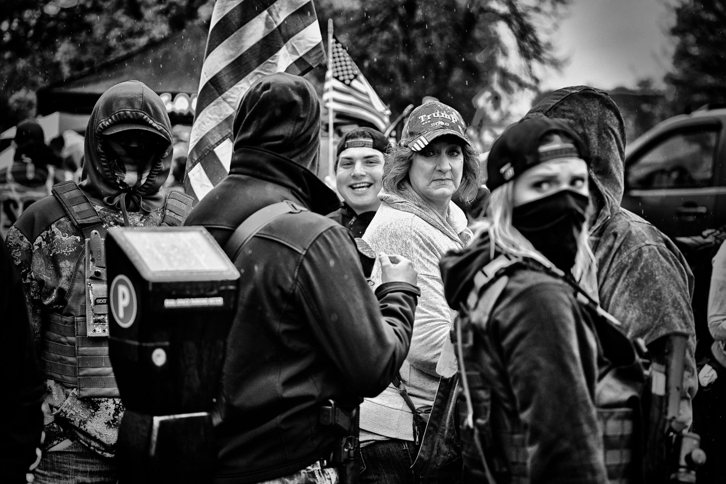 A group of protesters at an outdoor rally in rainy weather, some wearing Trump merchandise and others in tactical gear with face coverings. Some stare menacingly towards the camera. American flags are visible among the crowd.