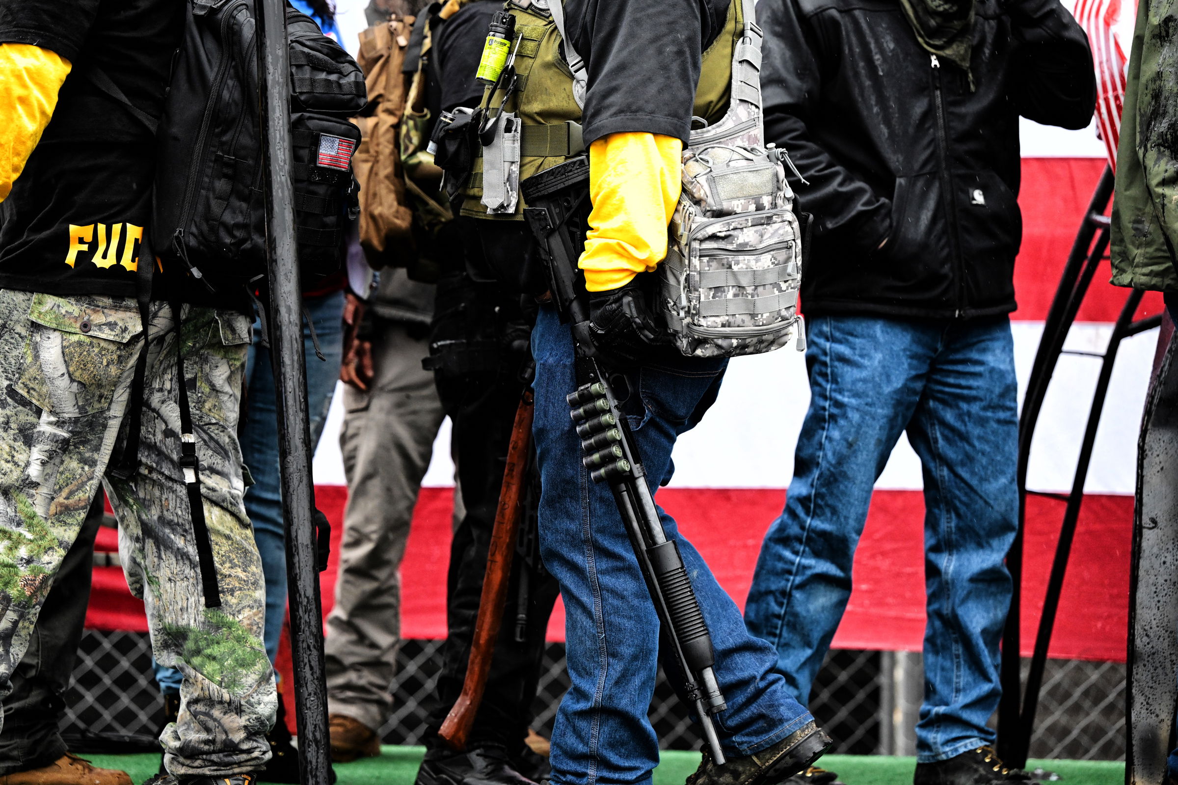 Close-up of armed protesters at a political rally, showing tactical vests, firearms, and yellow armbands. An American flag is partially visible in the background over a chain-link fence.