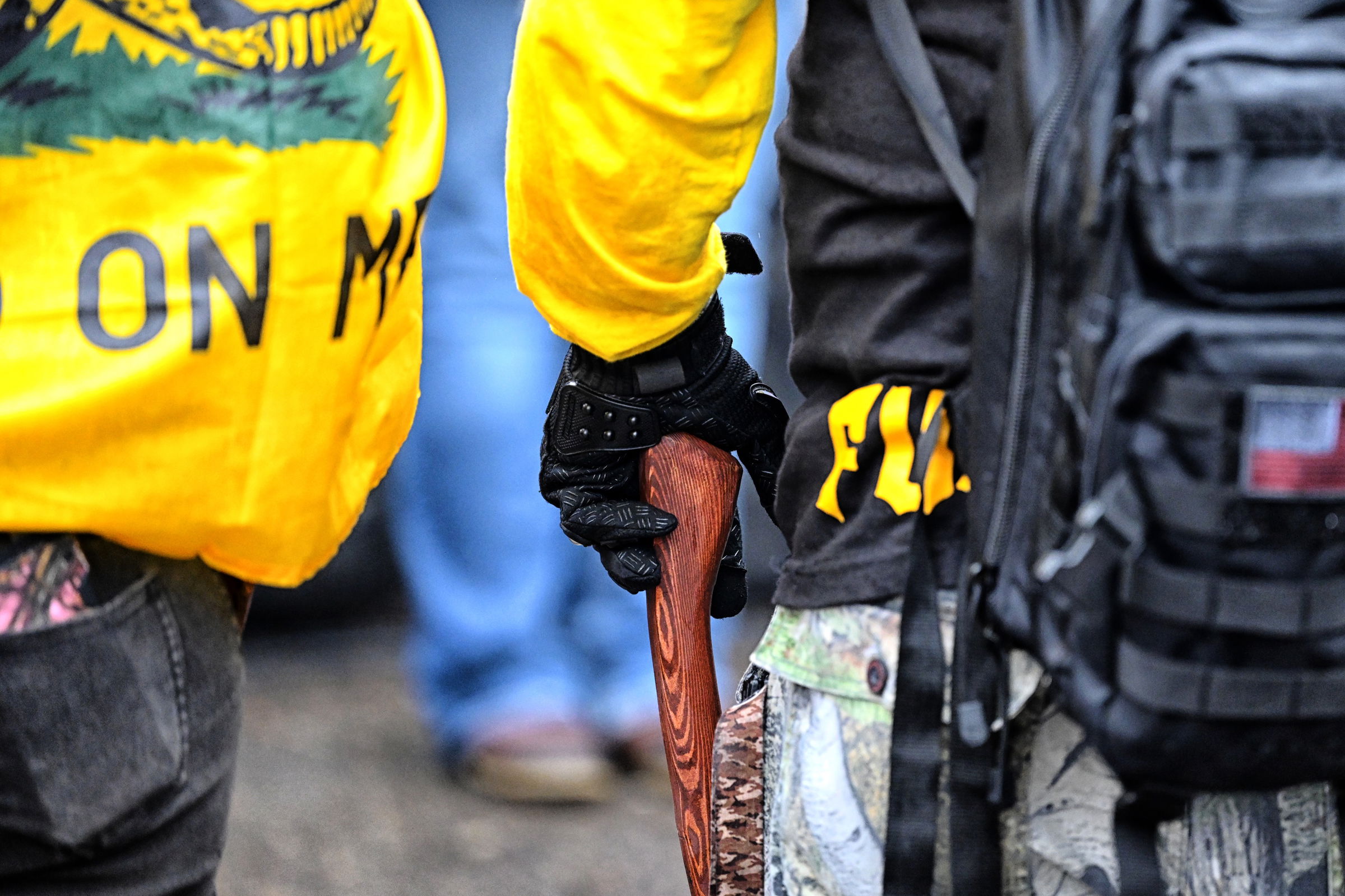 Close-up of two protesters wearing yellow garments, one wrapped in a "DON'T TREAD ON ME" flag, the other in tactical gear and a black shirt with the letters "FU" visible. One of the protester's gloved hands grasps a wooden axe handle.