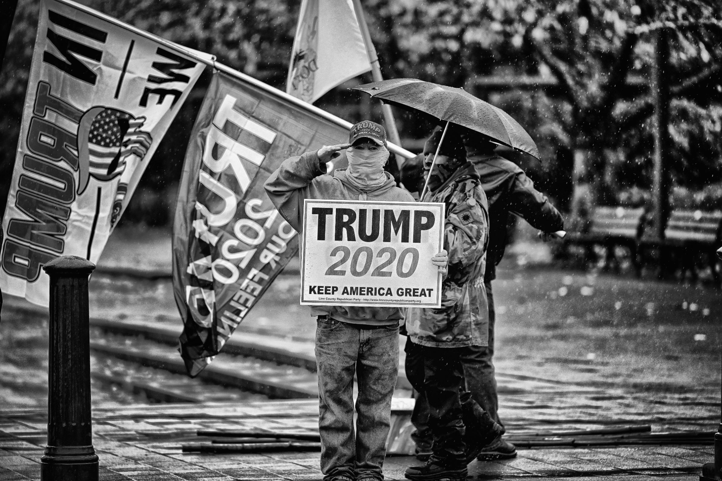 Trump supporters stand in the rain, one holding a "Trump 2020" sign and wearing a Trump hat while saluting the camera. Trump flags wave behind him.