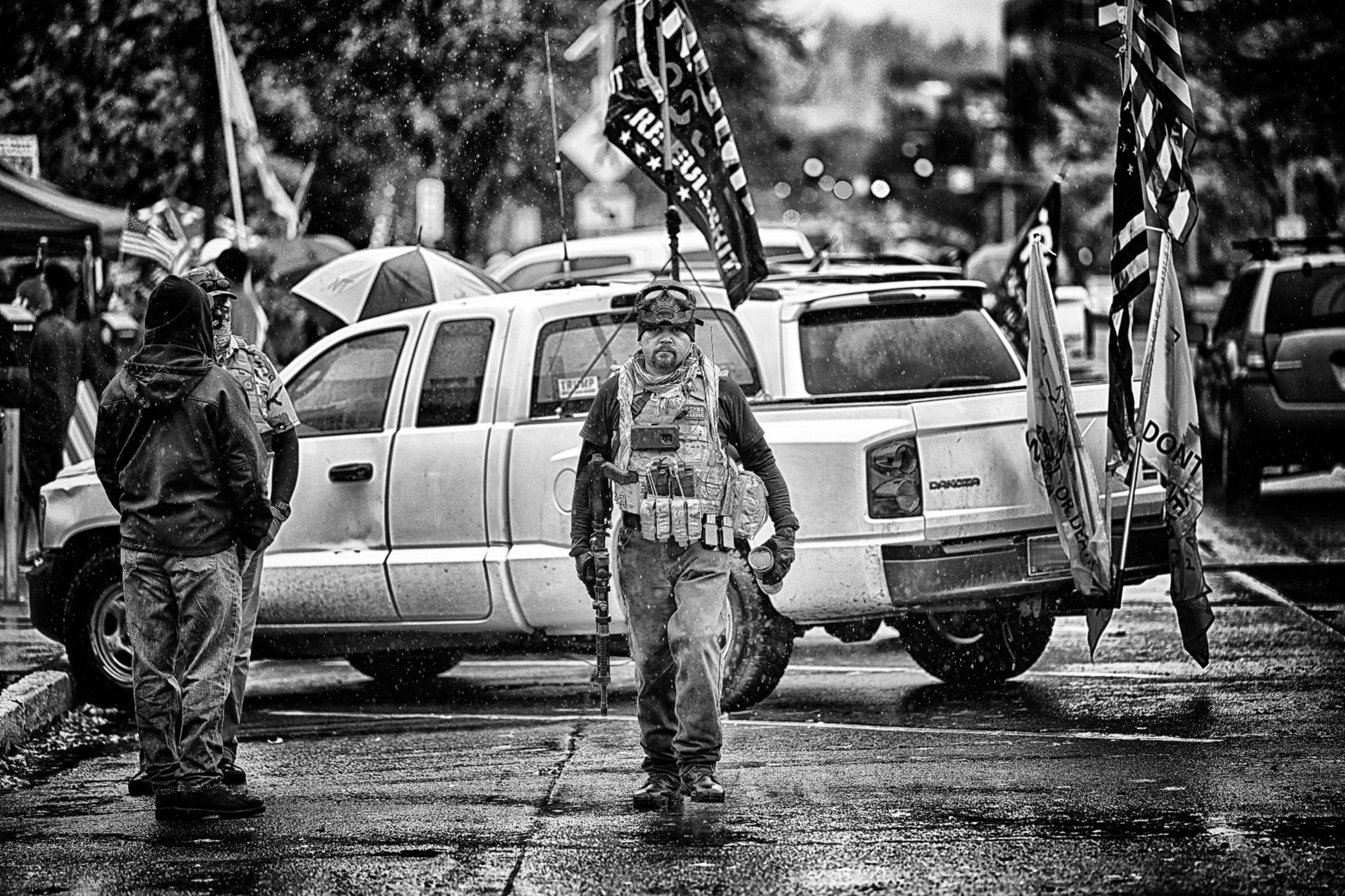 A rainy outdoor political rally. In the foreground, a man wearing military-style tactical gear and carrying a rifle is walking towards the photographer on the wet street, staring into the camera. Behind him is a white pickup truck with various American and Trump flags attached. Other individuals in rain gear can be seen nearby, and there are multiple vehicles and flags visible in the background.