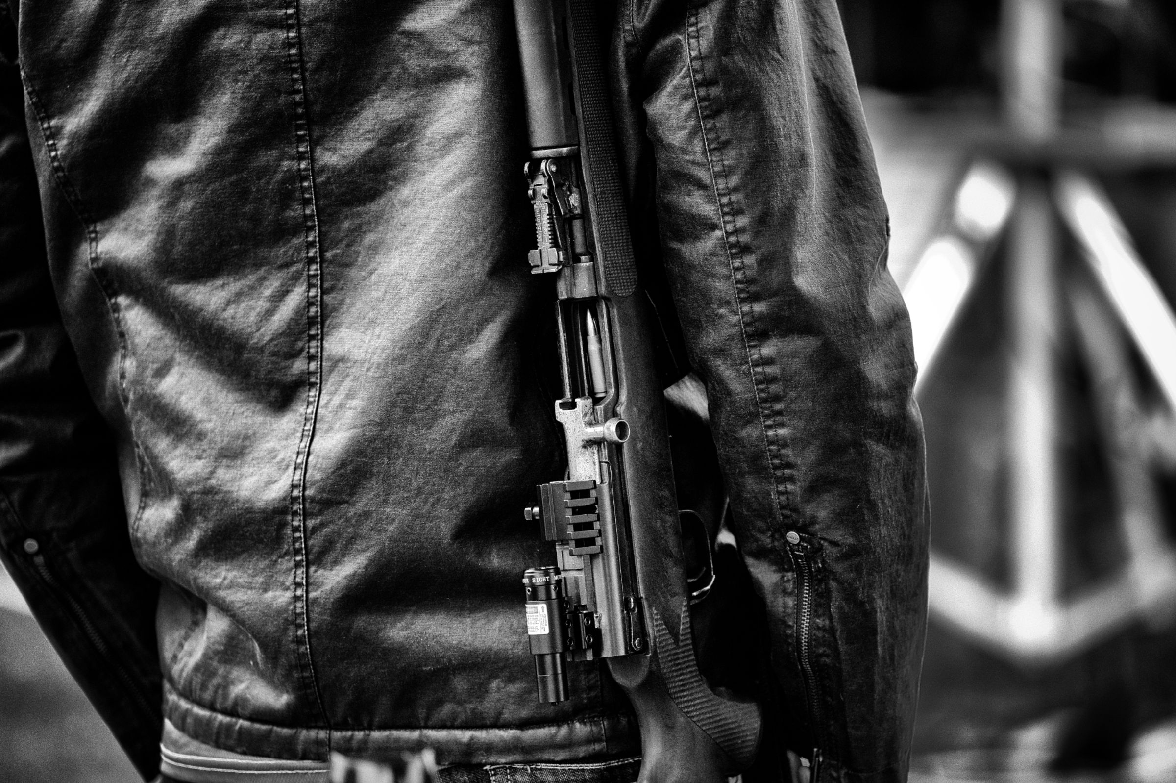 Close-up black and white image of a person wearing a leather jacket, with a semi-automatic rifle hanging at their side. A live round is visible in the chamber.