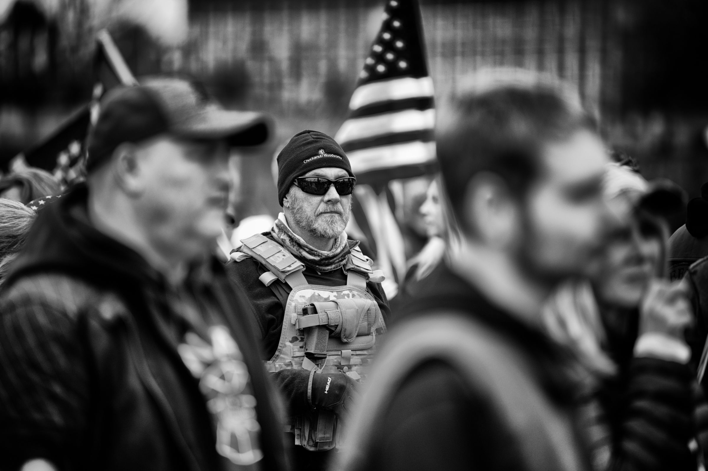 Black and white image of a bearded man in tactical gear and sunglasses standing amid a crowd at a political rally. He wears a knit cap and body armor with a handgun strapped to the front of his vest.