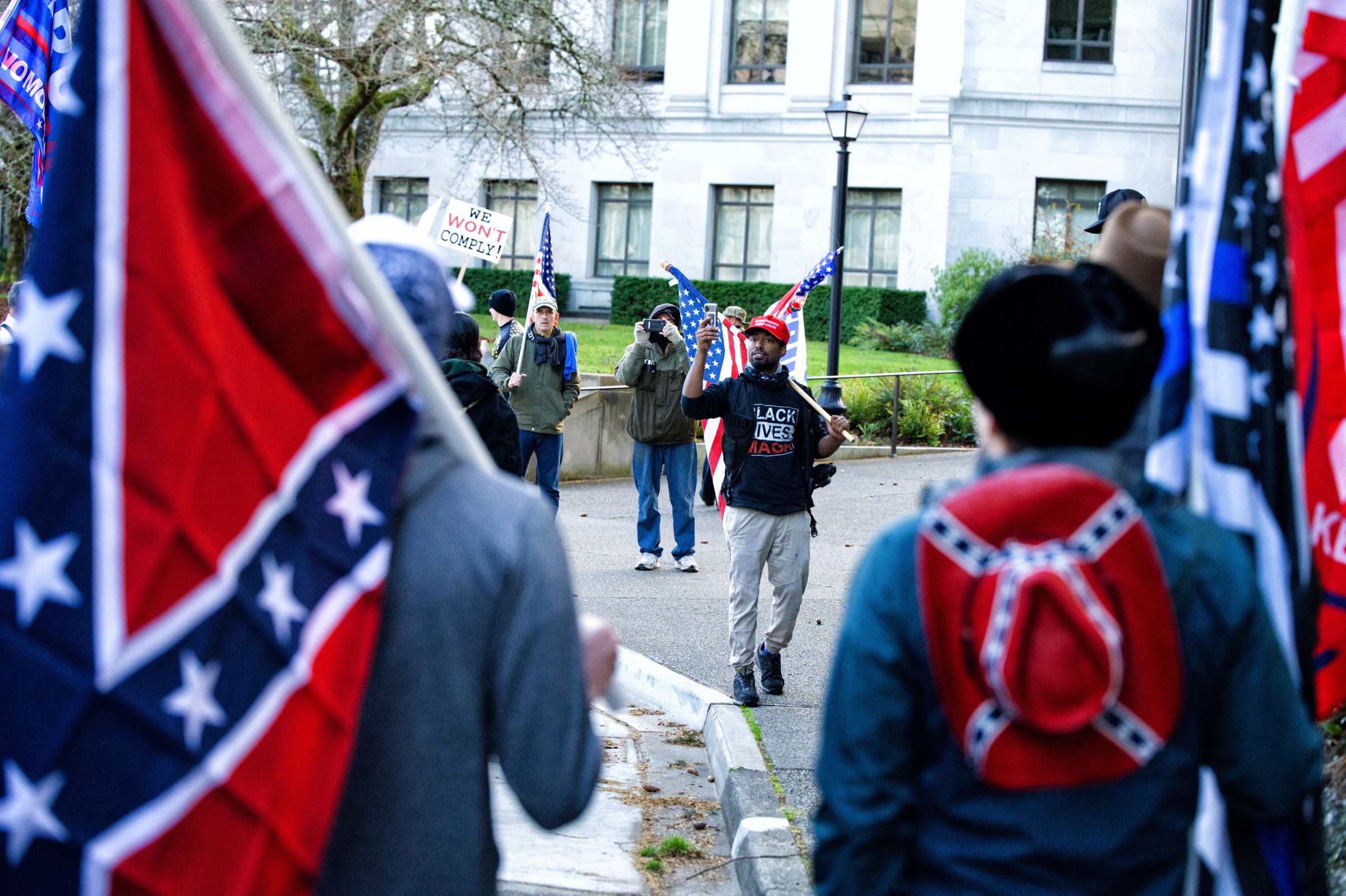 A political demonstration outside a government building. Two white men holding confederate flags are visible in the foreground. In the distance, a black man wearing a "BLACK LIVES MAGA" shirt and carrying an American flag faces the confederate flag carriers uncertainly.