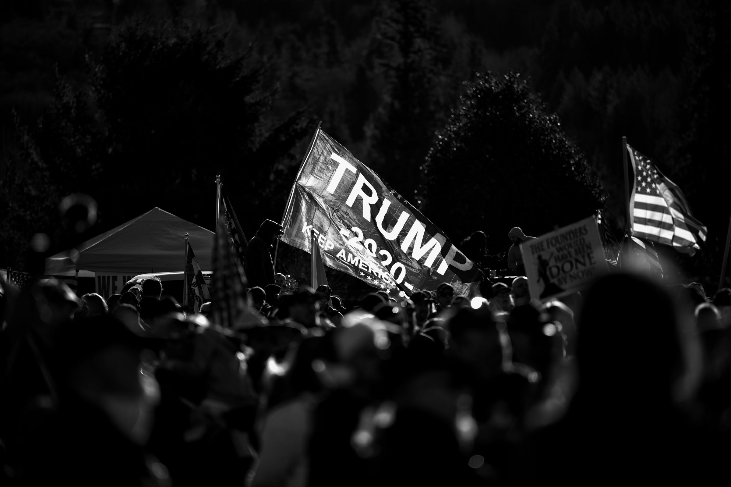 Black and white nighttime image of a crowded political rally, featuring a prominent "TRUMP 2020" flag among a sea of supporters. American flags and other signs are visible, illuminated against the dark background.