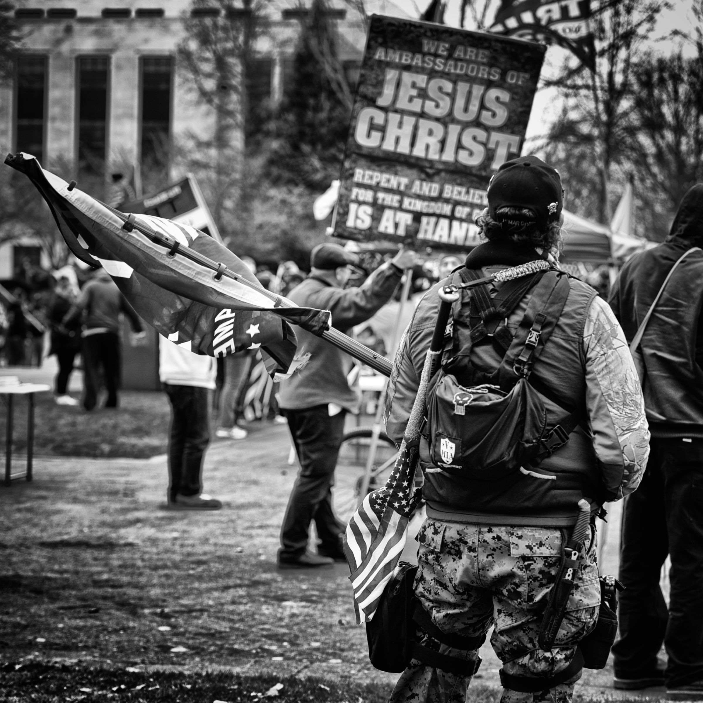 A man armed with a large knife and wearing camouflage clothing and tactical gear stands with his back to the camera, holding a Trump flag. Over his shoulder is slung a baseball bat with an American flag attached to it. A sign in the background reads "We are ambassadors of Jesus Christ."