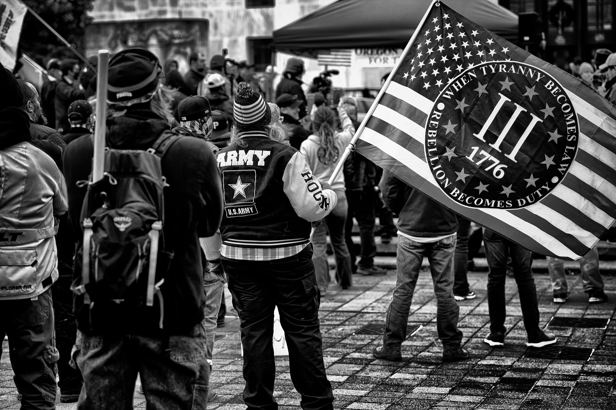 Demonstrators gather at a political event. A person wearing a U.S. Army jacket holds a modified American flag featuring the roman numeral III, the date 1776, and the text "WHEN TYRANNY BECOMES LAW REBELLION BECOMES DUTY".