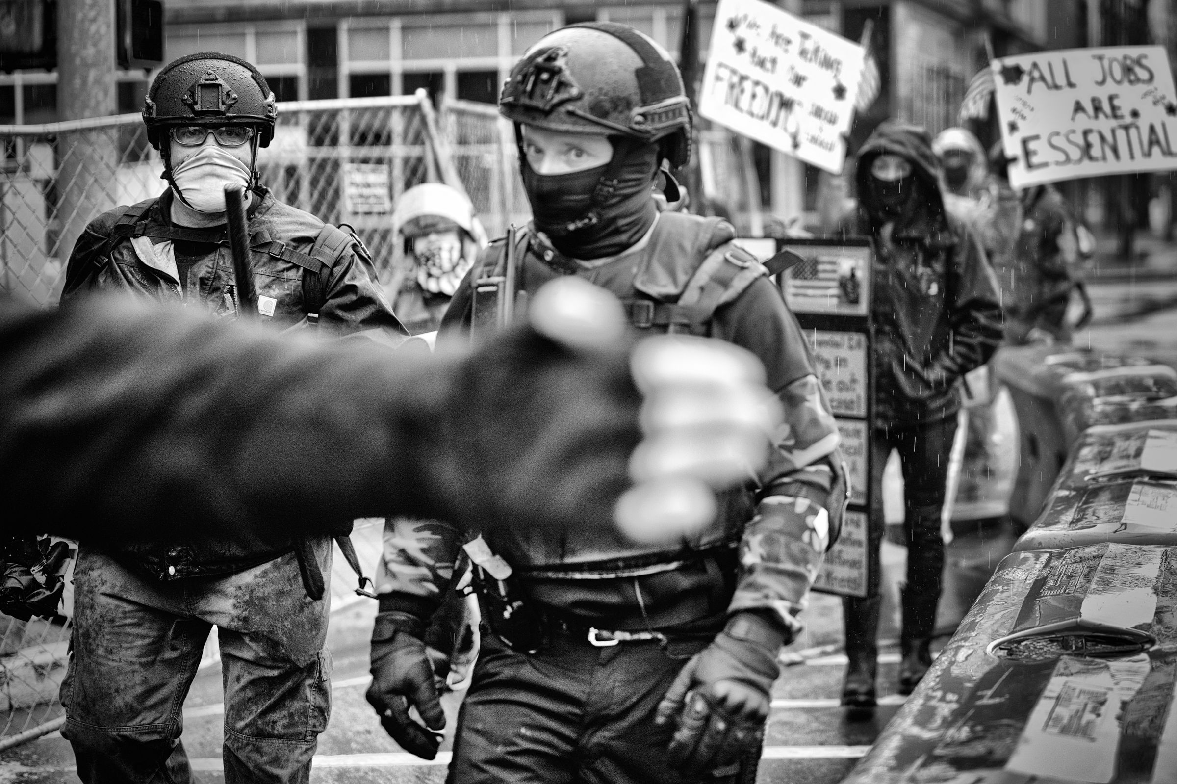 Two young men wearing military-style tactical helmets and gloves, both masked, stare menacingly towards the camera. One of the men brandishes a police baton. A third person's gloved hand obstructs the center of the camera.