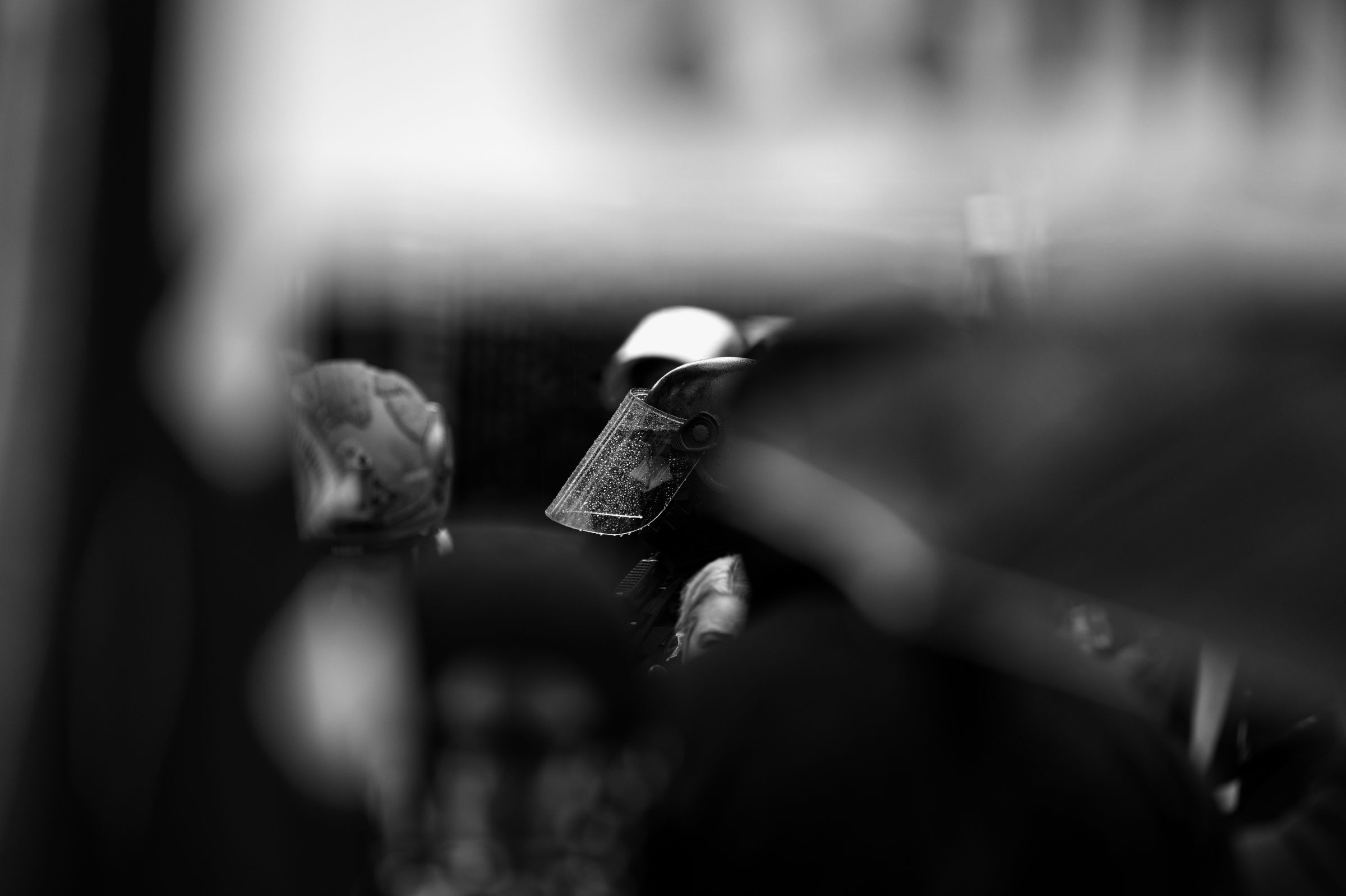 Close-up black and white photograph of a police officer's riot helmet with the face shield lowered amidst a crowd of protesters.