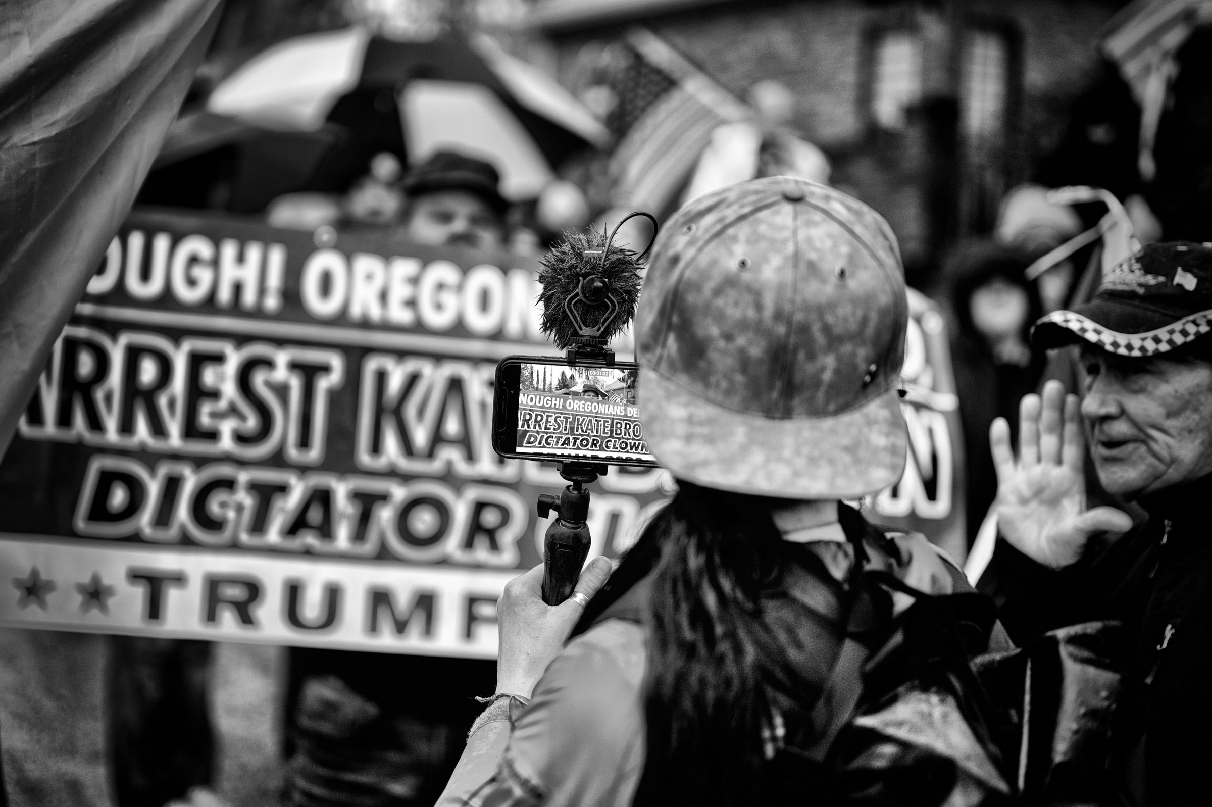 Black and white photo of a journalist holding a smart phone with a handheld monopod to record a crowd of protesters who are waving political signs and American flags. The journalist is in the foreground, with their back to the camera. A large banner can be seen in the background, with the focus of the picture on the camera screen where the partially obscured words "ARREST KATE BROWN DICTATOR CLOWN" are visible. A partially in-focus man stands to the right of the journalist with his hand raised to his mouth, taunting the journalist.