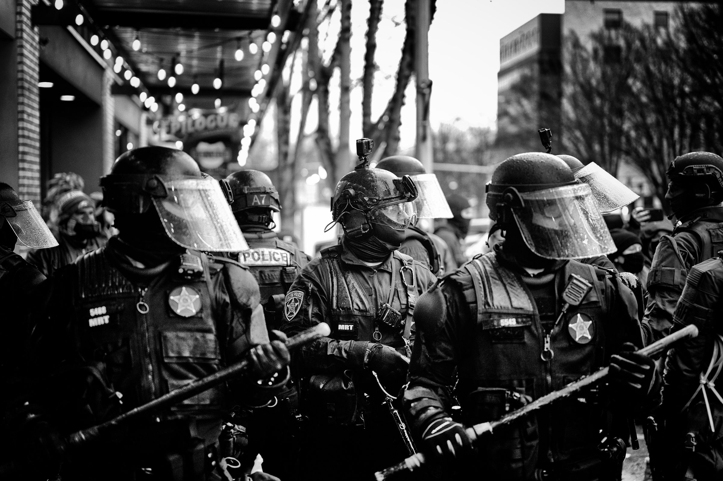 Helmeted police officers in riot gear brandish batons while standing in formation on a city street at night, illuminated by string lights overhead.