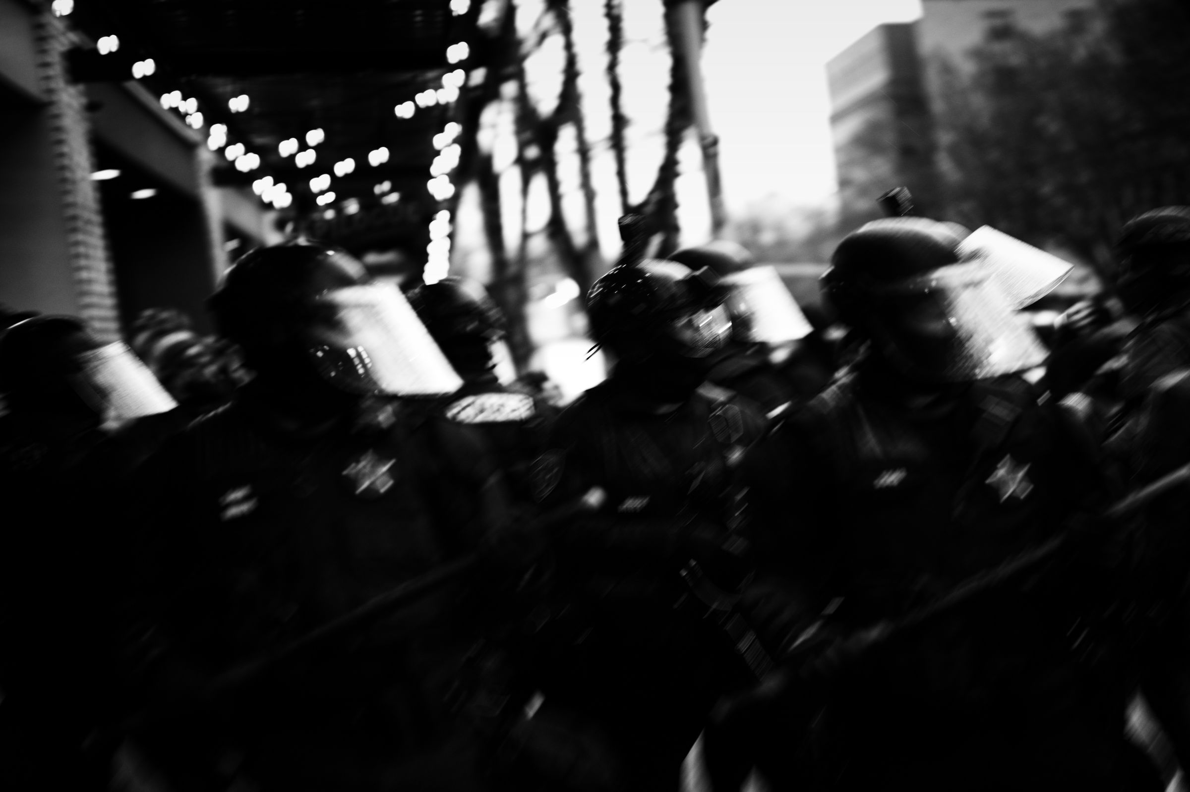 Blurred image of riot police in helmets and protective gear moving in formation, with string lights visible in the background.