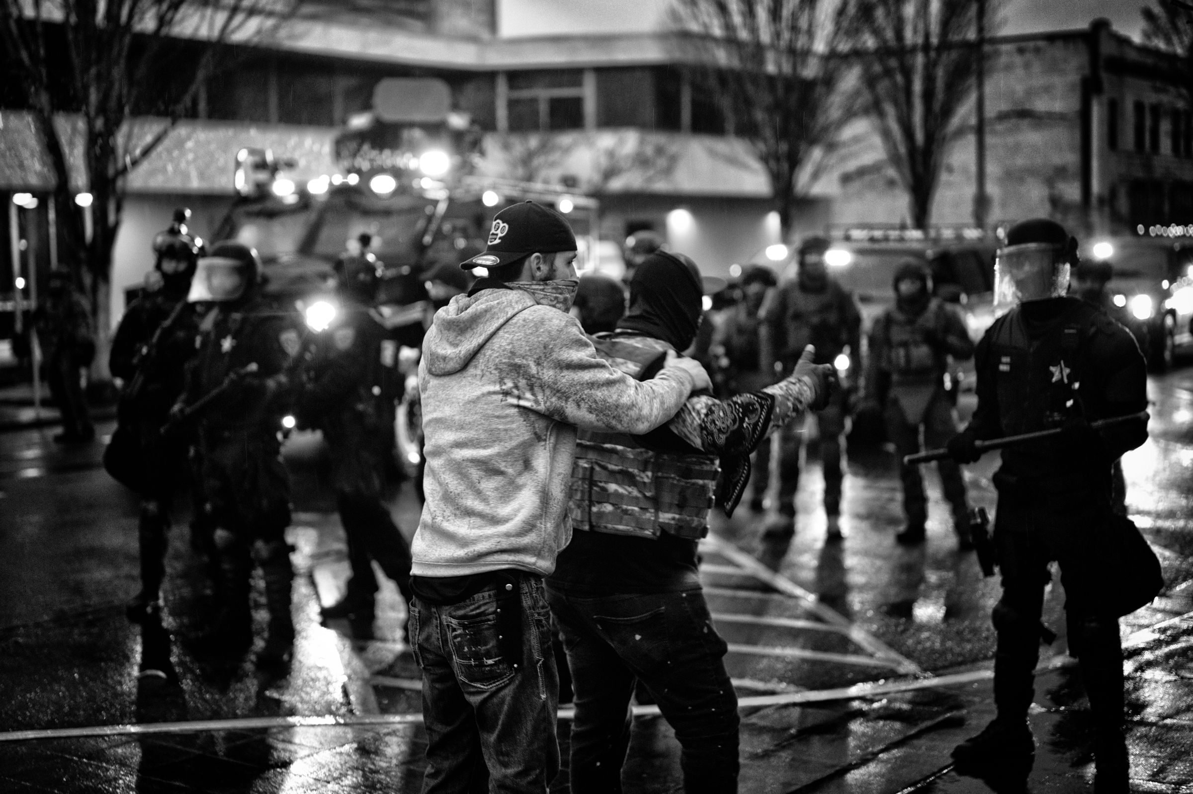 Two men, one wearing a hooded sweatshirt, the other a military-style armored plate carrier, face a group of advancing police in riot gear on a rainy city street at night. The man in the sweatshirt is attempting to hold back the man in the plate carrier as he gestures aggressively at the police.