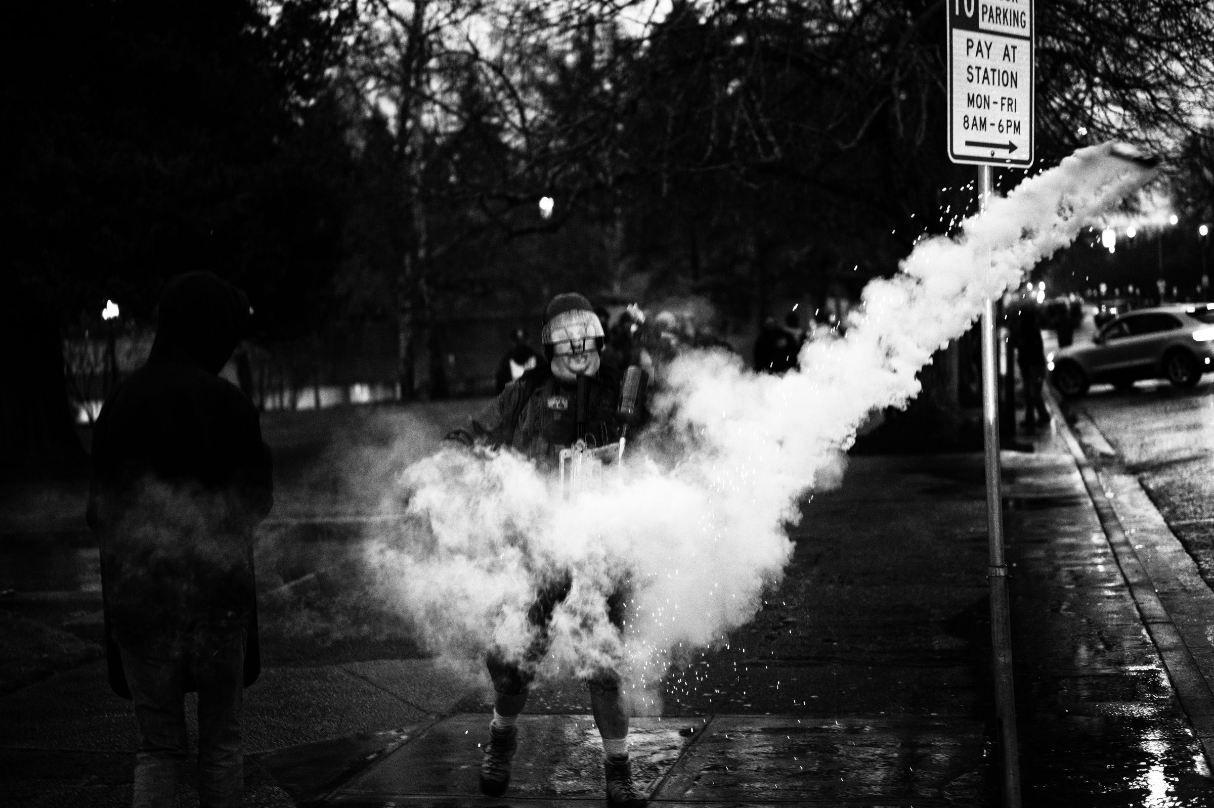 A cloud of smoke engulfs a man at the center of the frame, wearing an armored plate carrier and a protective helmet on a city sidewalk at night. The smoke trails behind a thrown canister frozen mid-flight to the right of a no parking sign on the right-side of the frame.