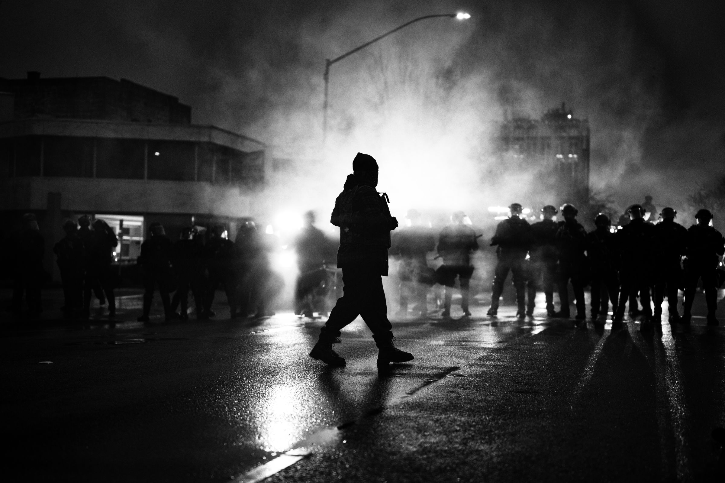 Black and white silhouette of a lone person on a wet street at night, facing a line of police in riot gear partially visible in the background amid smoke or tear gas.