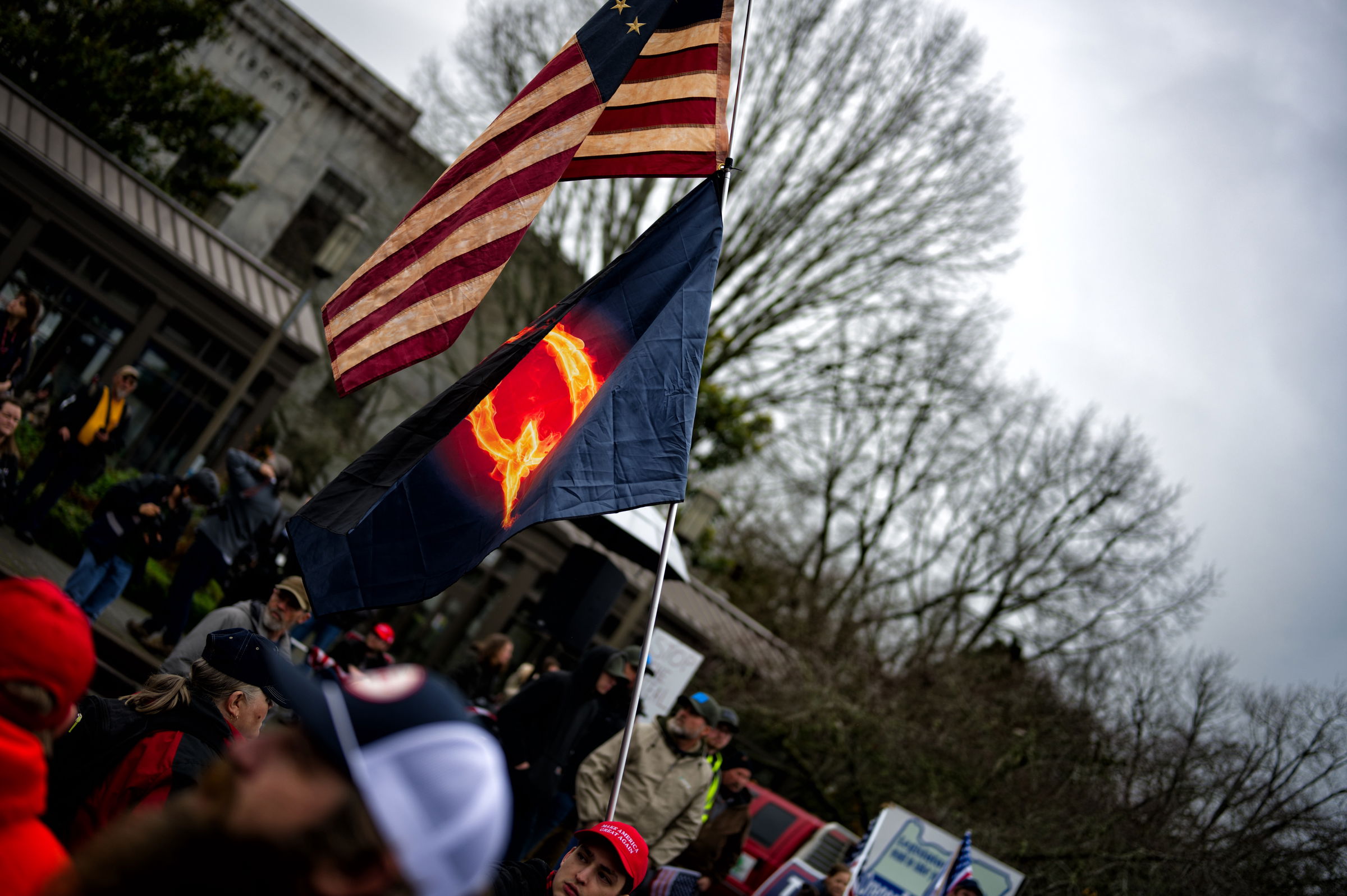 A man in a MAGA hat holds a large American flag and QAnon flag at political rally with crowd visible in background. The Q graphic on the QAnon flag appears to be engulfed in flames.