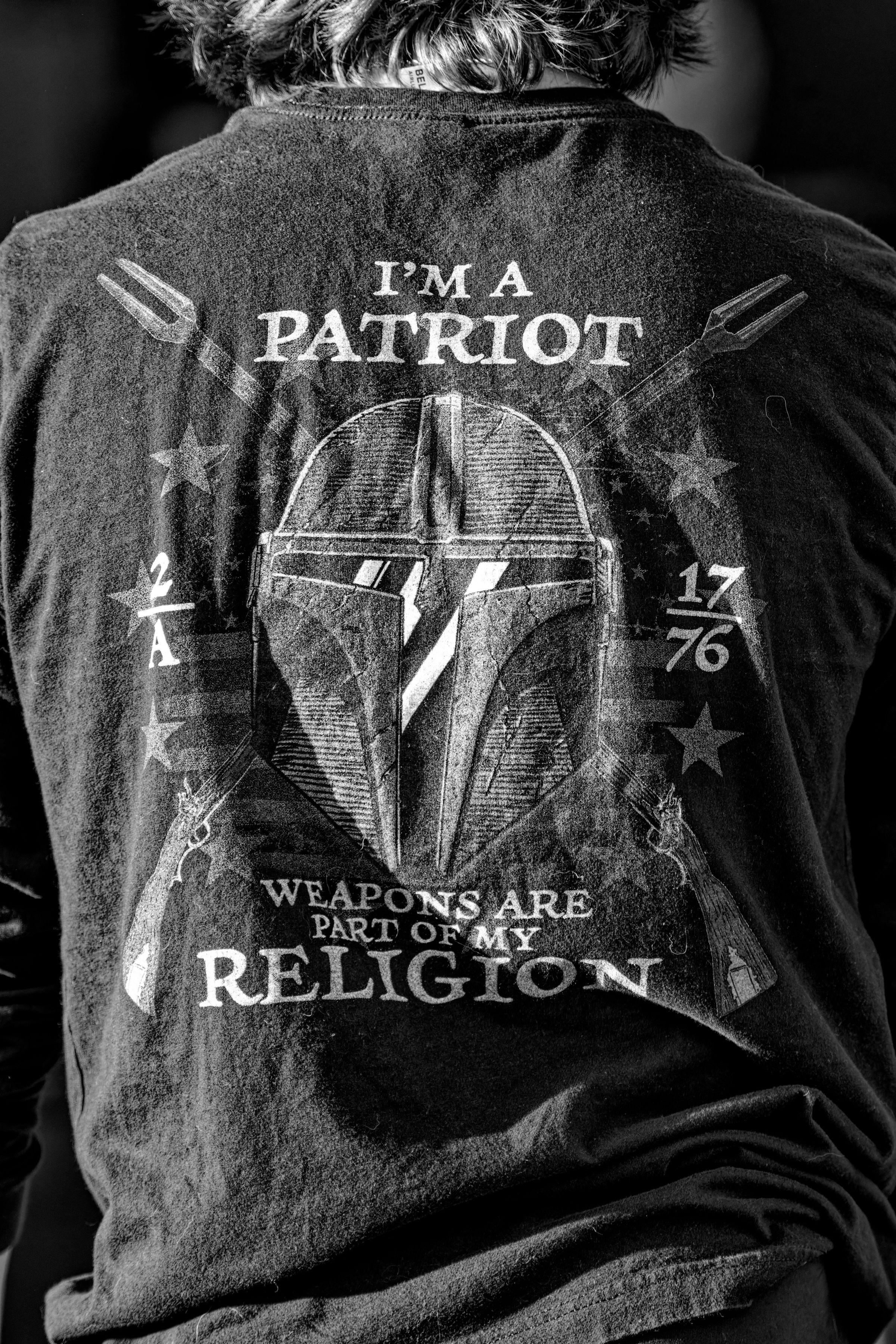 A black and white photo of a young man standing with his back to the camera. His dark t-shirt reads, "I'M A PATRIOT, WEAPONS ARE PART OF MY RELIGION". The text surrounds a gladiator-style helmet, muskets, and 2/A and 17/76 iconography.