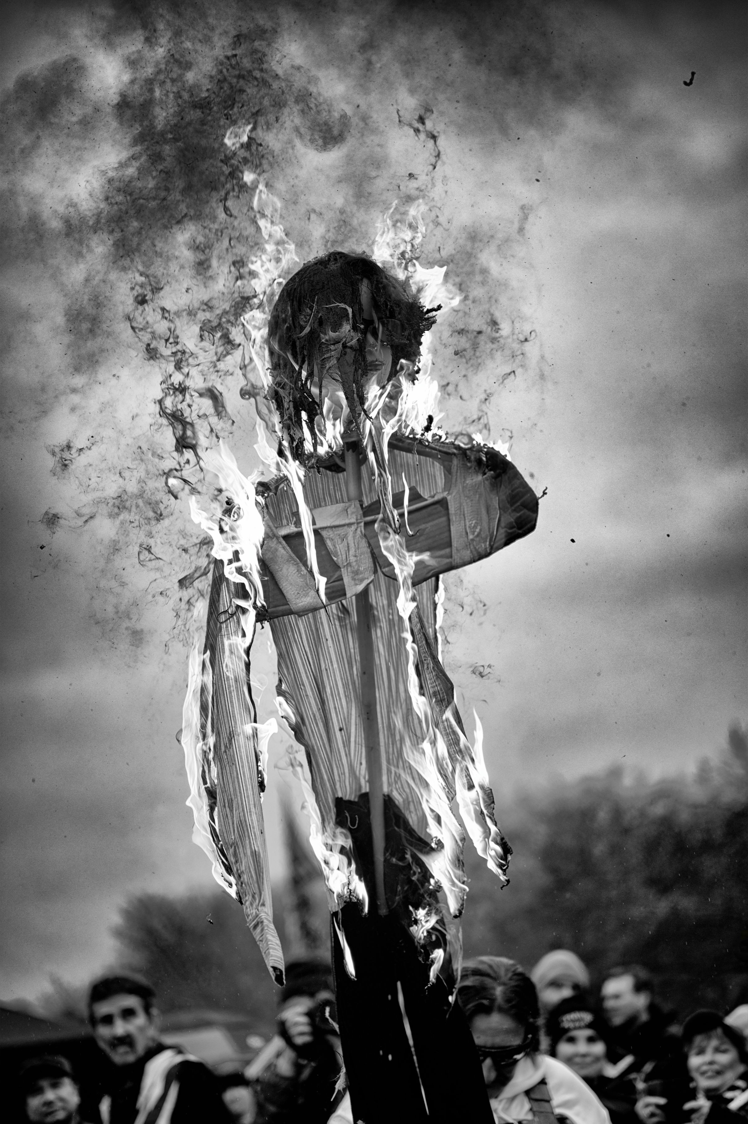 A black and white photo of a burning effigy engulfed in flames and dark smoke, held aloft with grinning onlookers in the background.