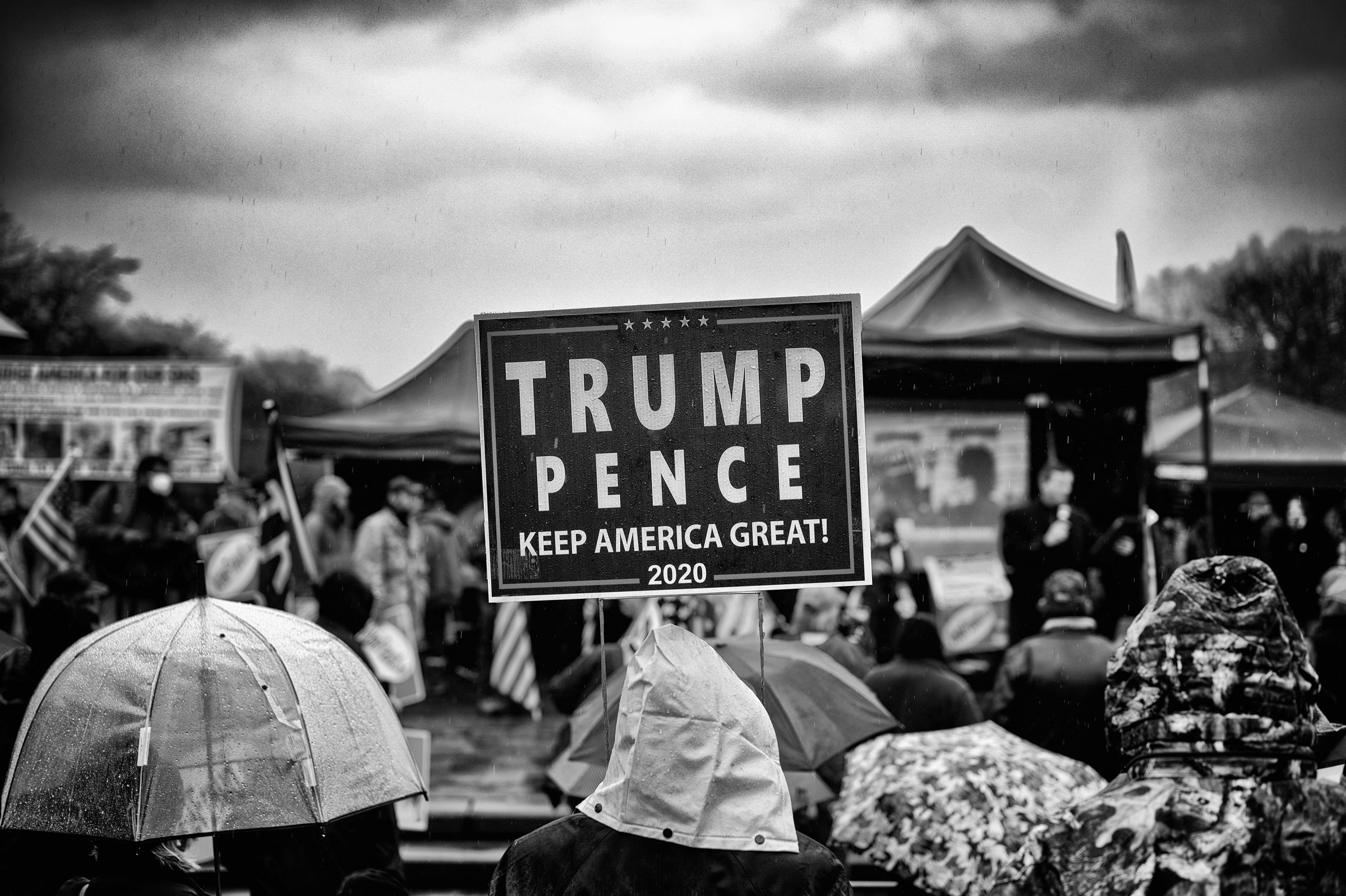 A person holds a campaign sign that reads "TRUMP PENCE KEEP AMERICA GREAT! 2020" at a rainy outdoor political rally, amidst a crowd of people in raincoats and holding umbrellas.