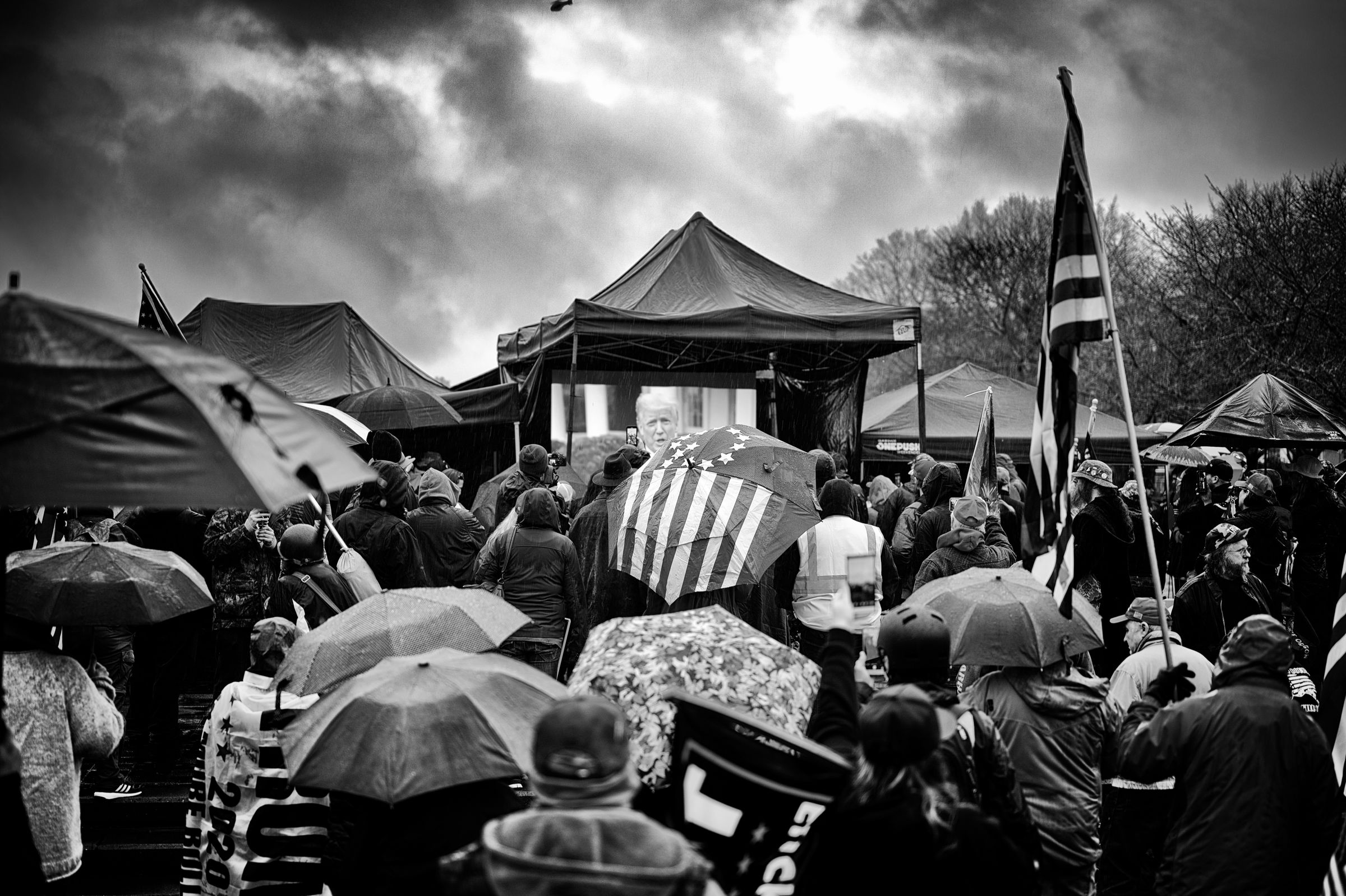 A large crowd with raincoats and umbrellas gathers around tents at a political rally on a rainy day in Oregon, with an American flag-themed umbrella in the foreground. In the background, President Donald Trump is visible on a TV screen, addressing protesters at the U.S. Capitol. A news helicopter hovers high overhead.