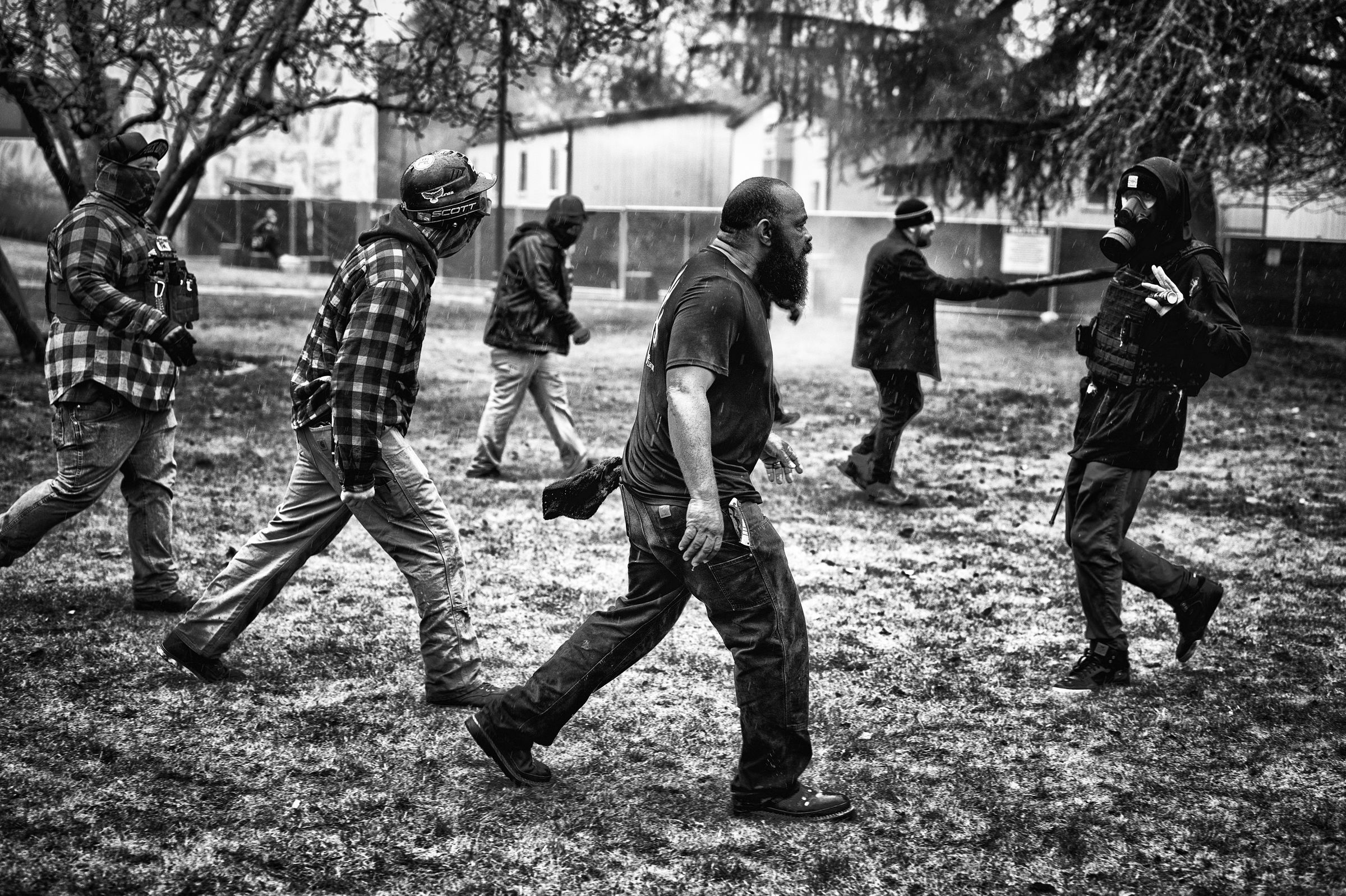 A group of men, some wearing tactical vests and protective helmets, walk across a grassy area, approaching an individual wearing a gas mask who is walking backwards with their hands partially raised. A large folding knife is visible, attached to one of the advancing men's pockets.