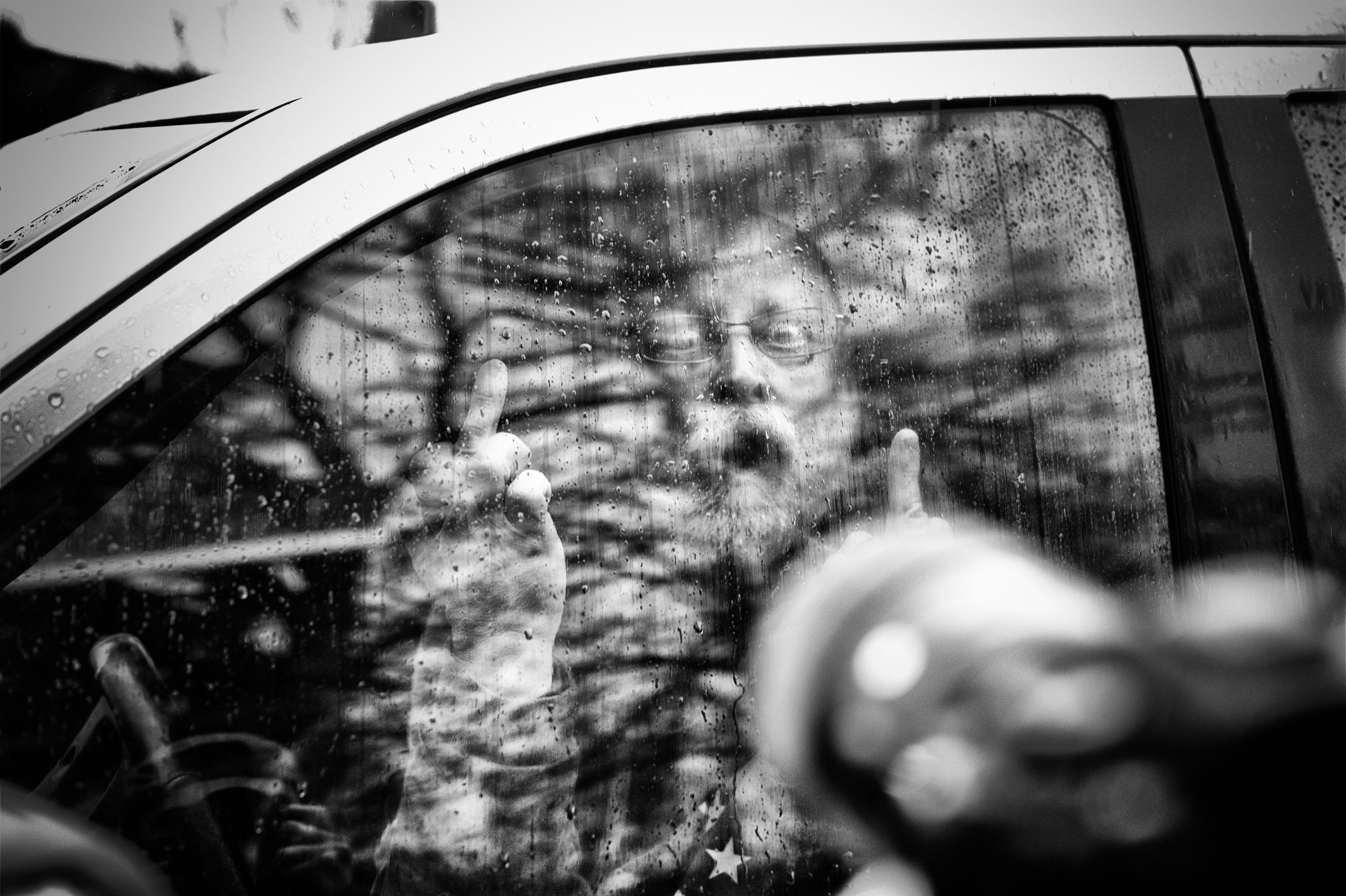 A close-up black and white photo through a rain-spattered car window shows an older man with a beard gesturing wildly with his middle fingers at a second camera in the foreground. The man's face is partially obscured by reflections on the glass.