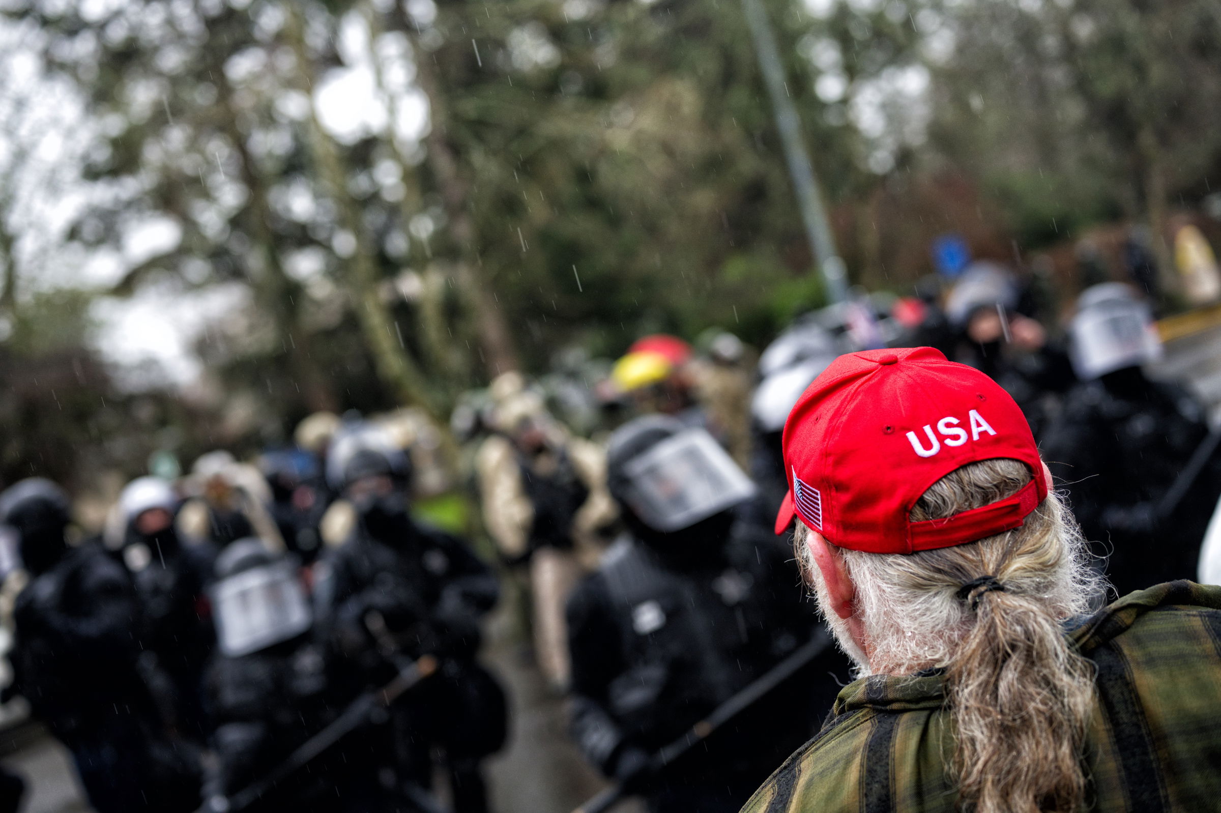 A man wearing a plaid shirt and a red "USA" cap stands facing a line of law enforcement officers in riot gear.