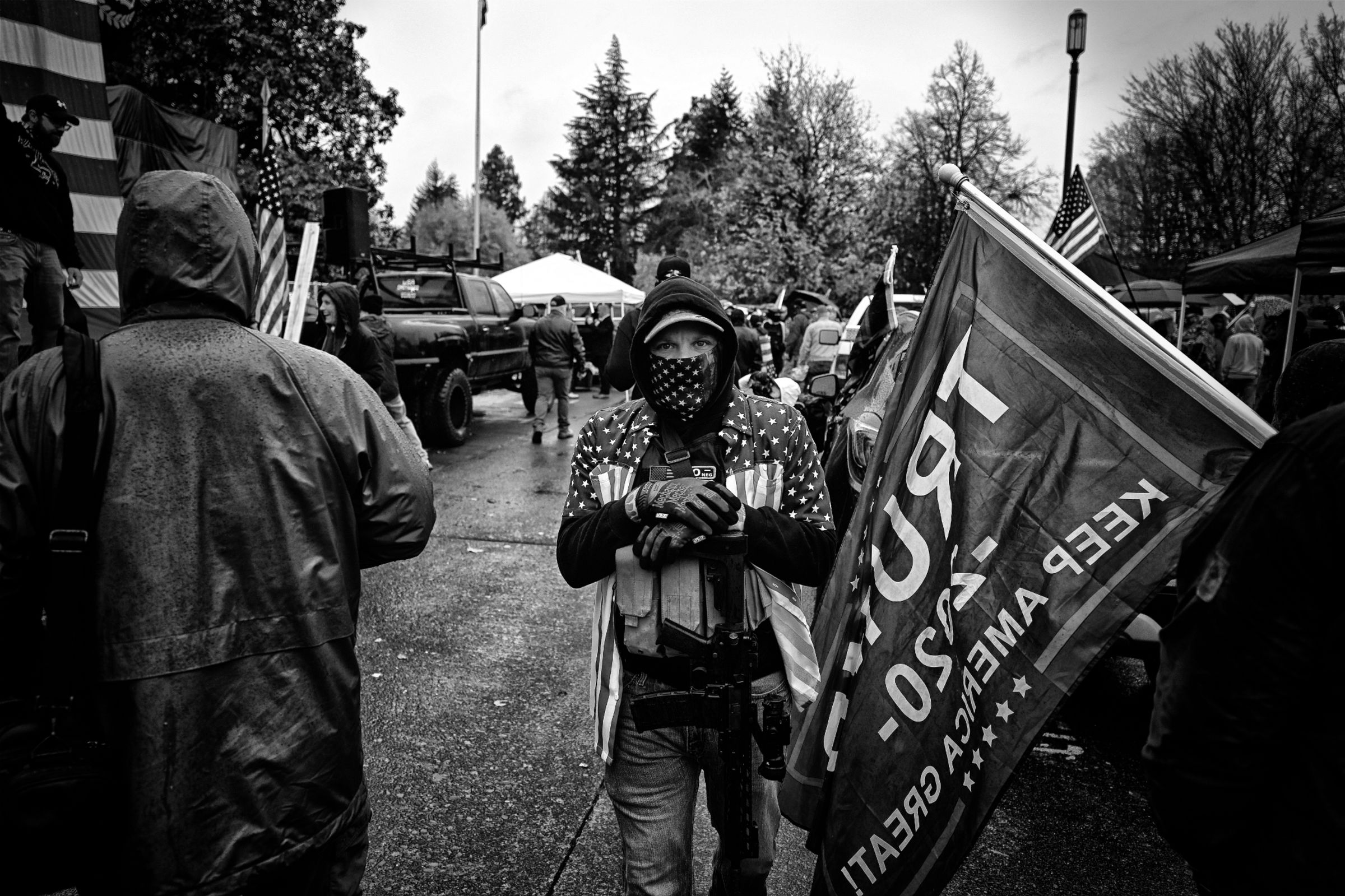 A black and white photo of a man at a crowded political rally. He's surrounded by American flags and a "Trump 2020" flag to his right. The man wears an American flag shirt over a military-style armored plate carrier with several magazine pouches attached to the front. An AR-style rifle with a magazine and scope is slung over his right shoulder and hangs vertically in front of his torso, with his hands resting on the stock of the rifle. The man's lower face is hidden by an American flag mask that matches his shirt. His eyes look unflinchingly at the camera.