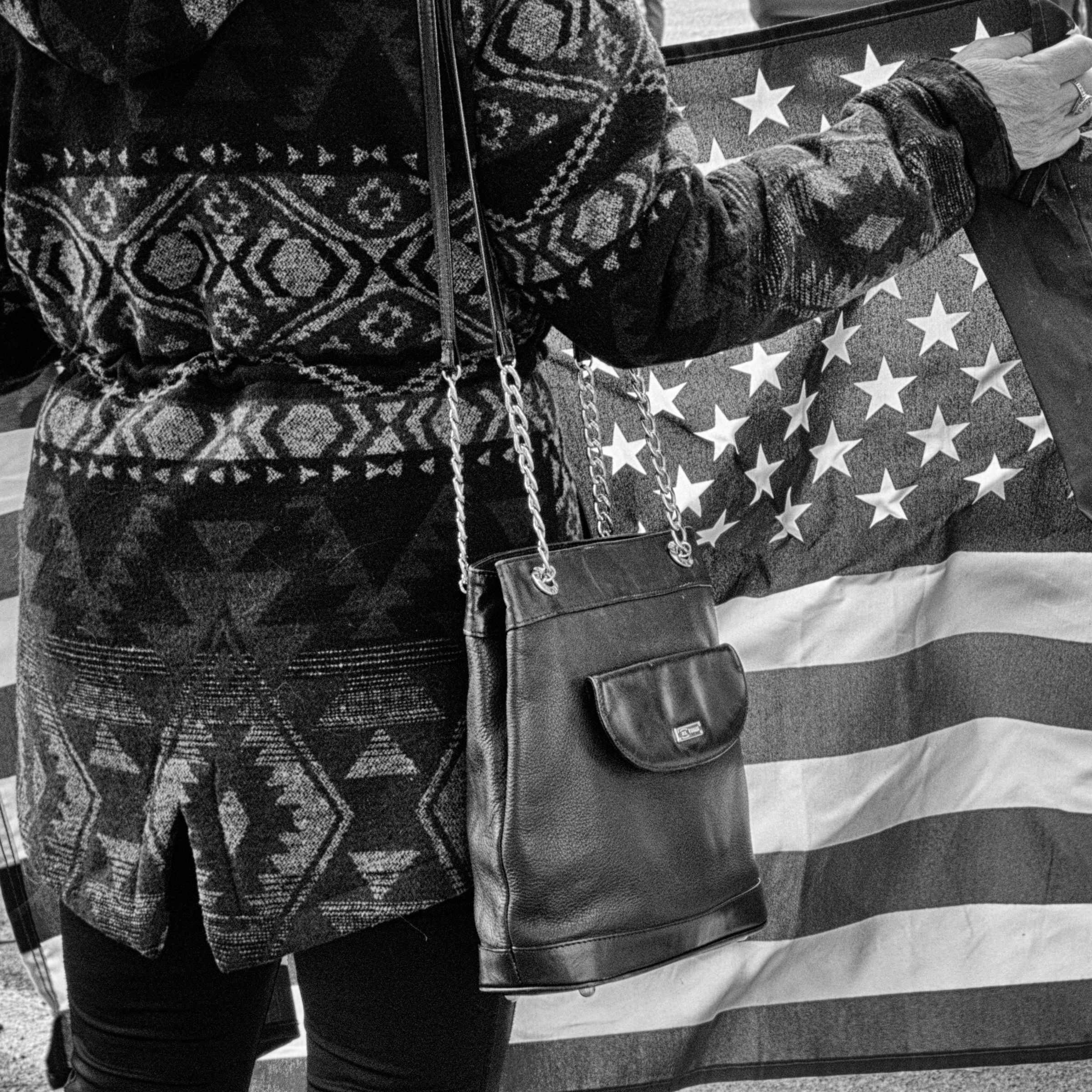 A person wearing a patterned sweater and carrying a leather handbag stands in front of an American flag.