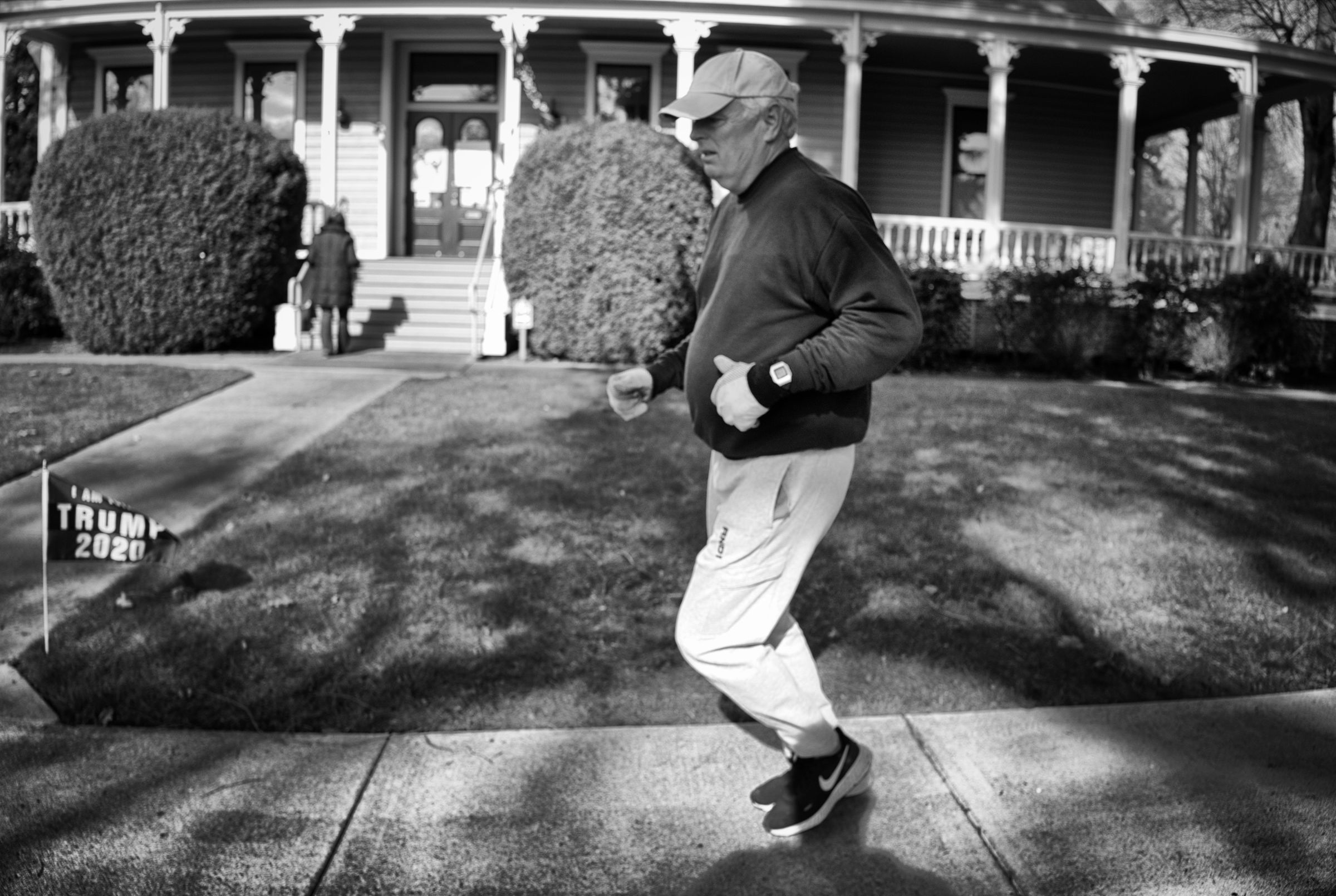 An older man in casual athletic wear jogs past a house with a wraparound porch, with a small "Trump 2020" yard flag visible in the background